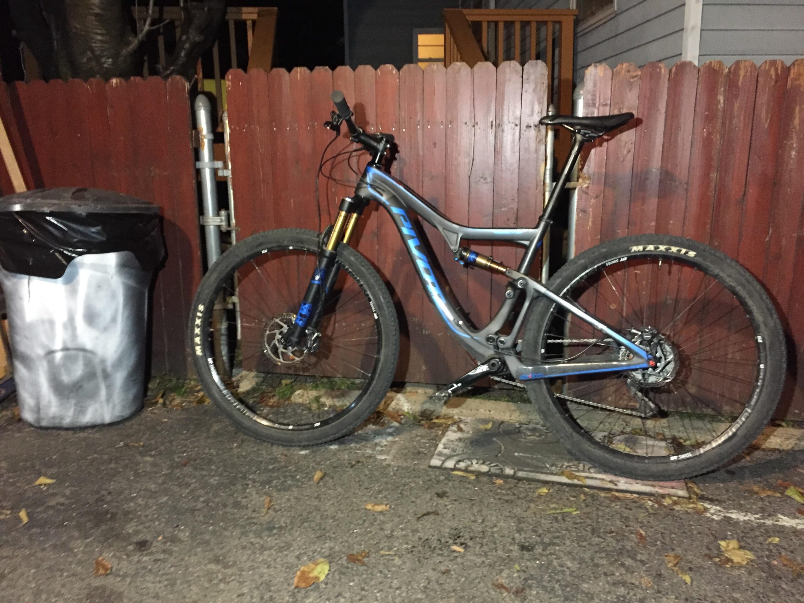 Pivot Mach 429SL Carbon: A mountain bike leaning against a wooden fence in an alley, next to a trash can. The bike has a blue and silver frame with large tires, and the surrounding area shows a paved ground scattered with leaves. Dim ambient lighting indicates it is evening.