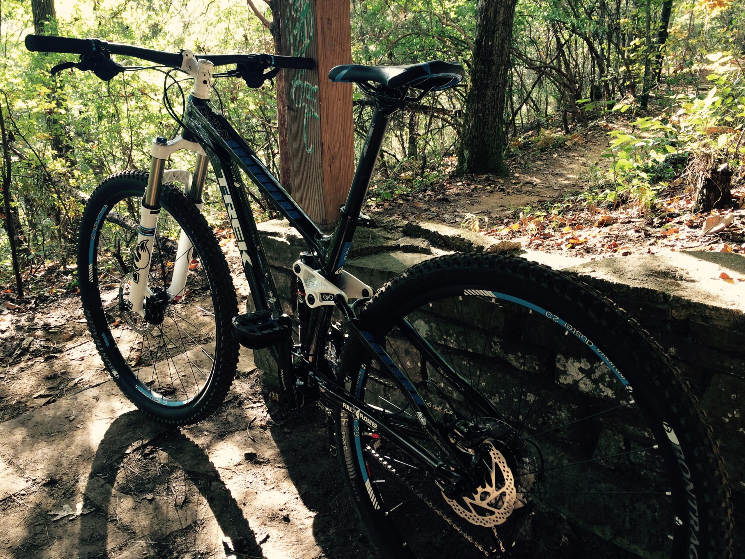 Trek Fuel EX 7 29: A mountain bike resting against a stone wall on a wooded trail, with dappled sunlight filtering through the trees, showcasing the bike’s detailed design and rugged tires.