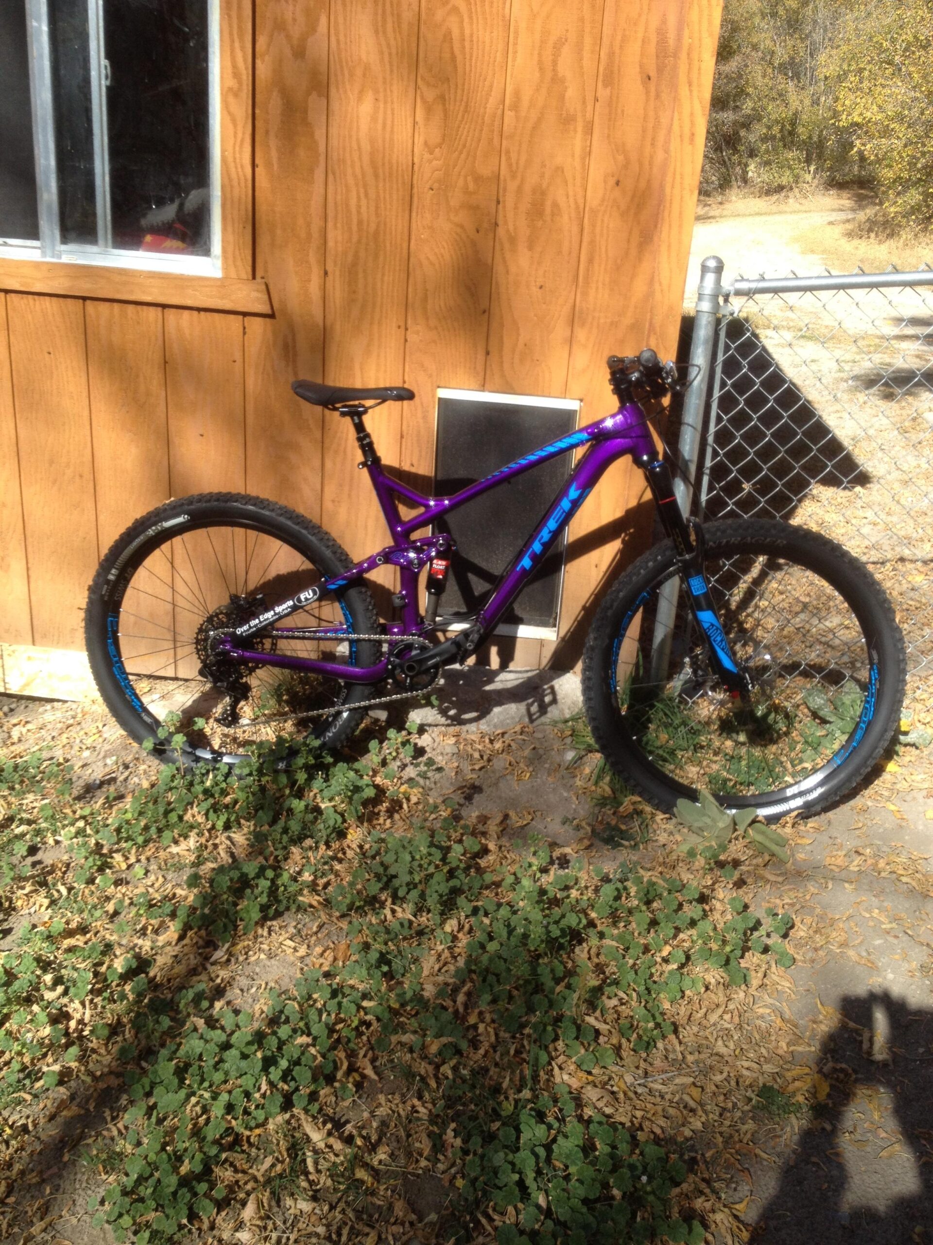 Trek Remedy 9: A purple and blue mountain bike leaning against a wooden wall, surrounded by patches of green plants and fallen leaves. A window and a fence are in the background, with a dirt path visible beyond the fence.