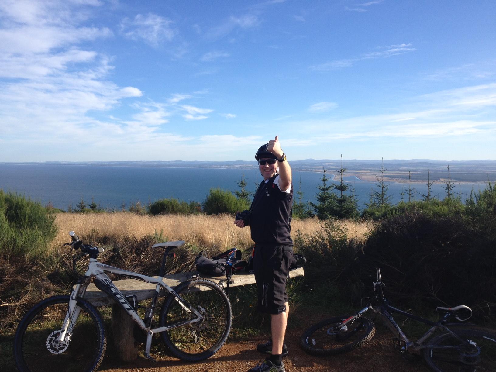 A person with a helmet and sunglasses gives a thumbs-up while standing next to two mountain bikes parked on a scenic overlook. The background features a view of a blue sea and distant hills under a partly cloudy sky. Learnie Red Rocks mountain bike trail.