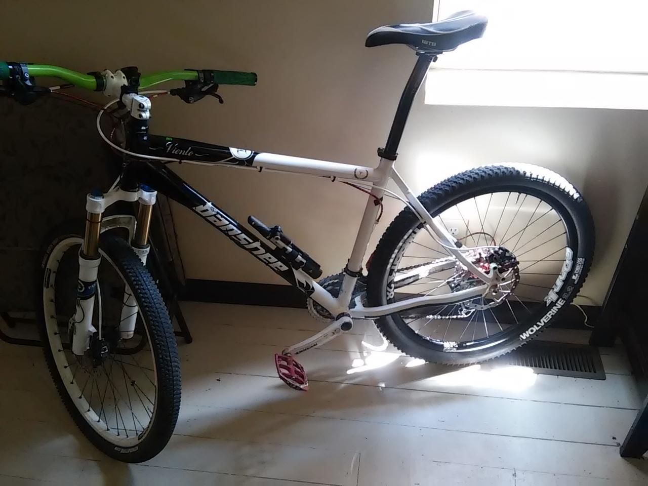 Banshee Viento: A mountain bike with a black and white frame, featuring green handlebars and pink pedals, is positioned in a sunlit room. The bike has noticeable front suspension forks and wide tires, and it is leaning against a wall near a window.