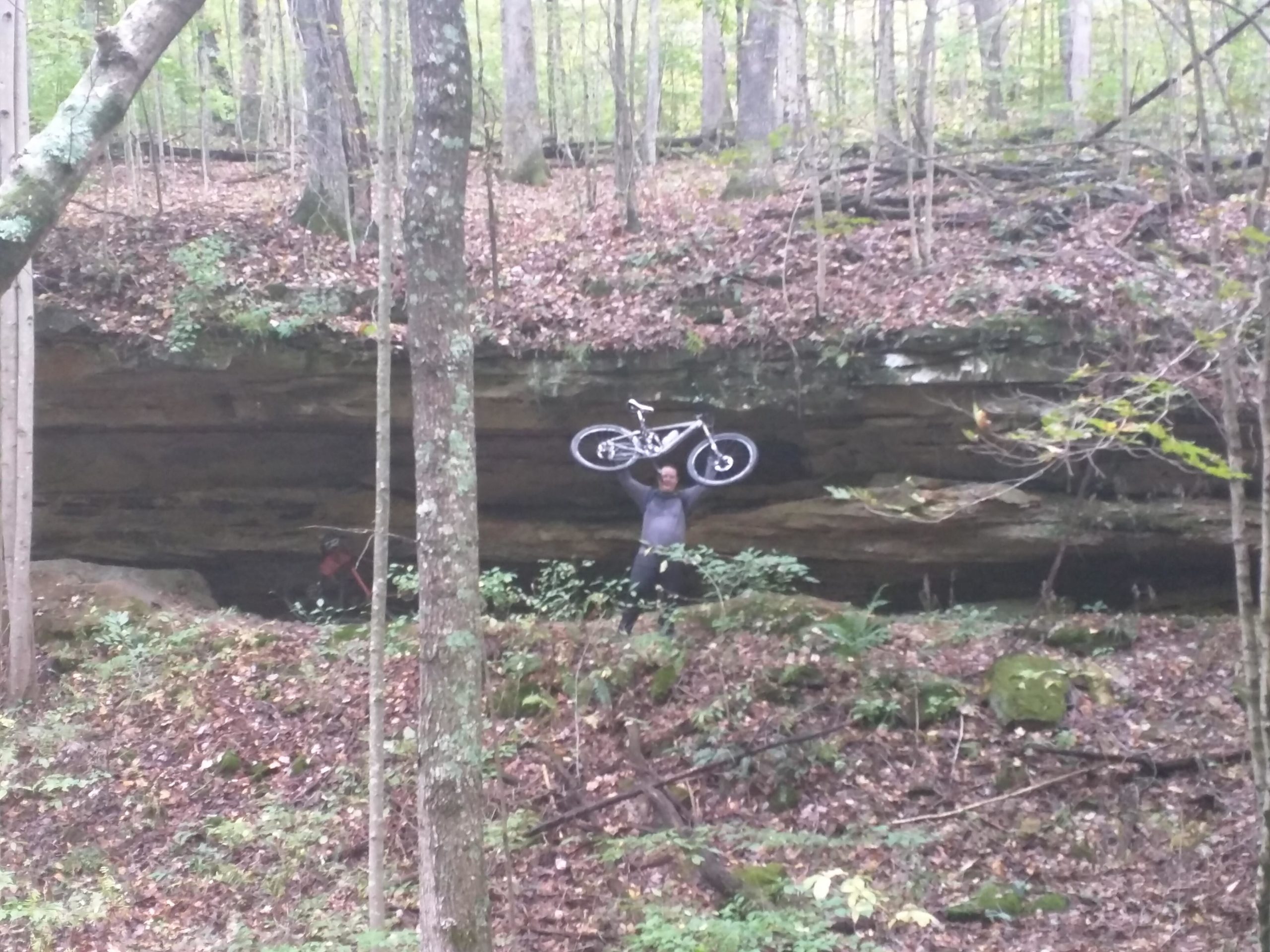 A person holding a bicycle above their head stands in a forested area with rocky terrain. The background features large stone formations, surrounded by trees and fallen leaves. Buffalo Trace mountain bike trail.