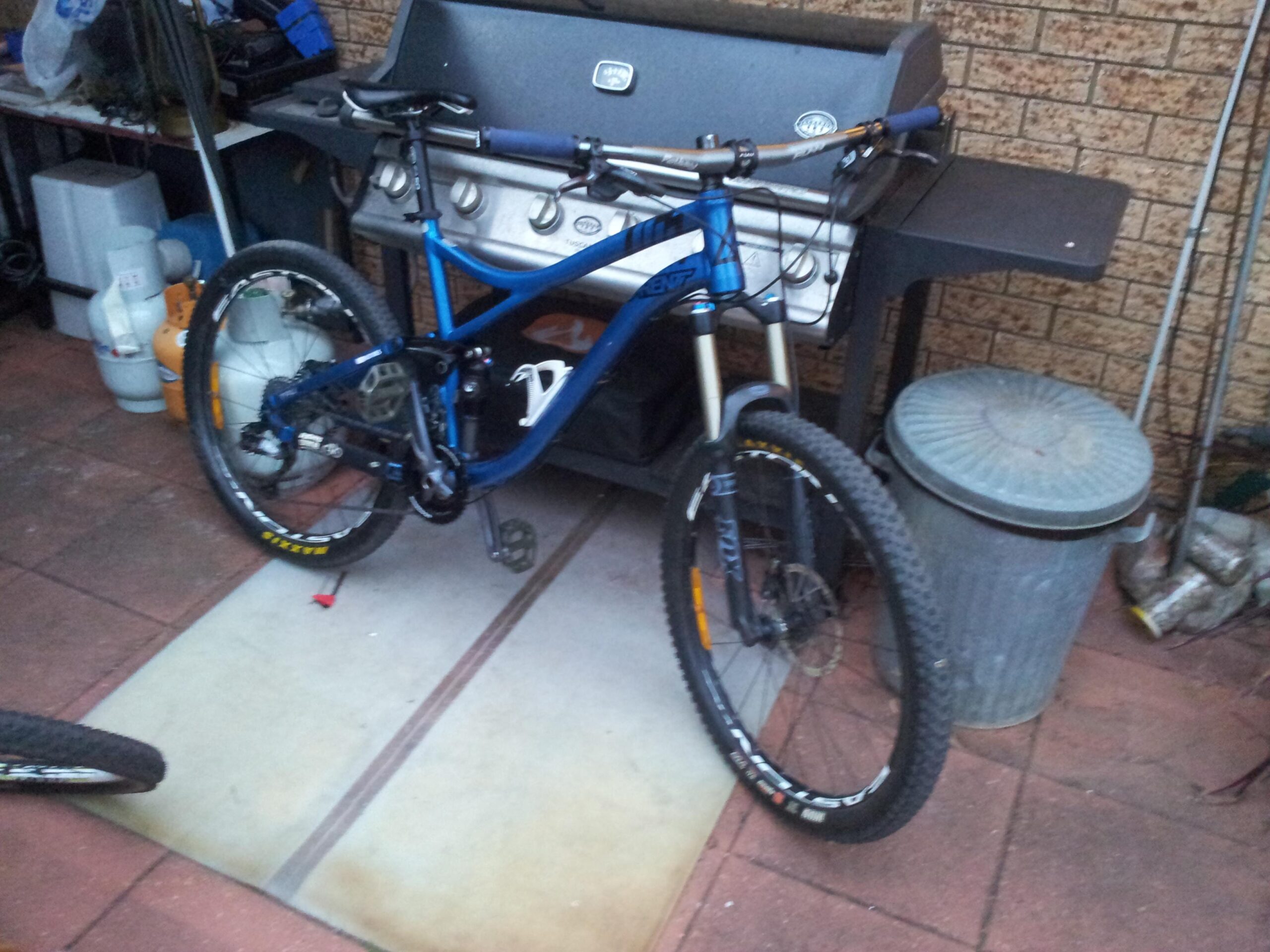 Avanti Avanti Torrent full suspension mountain bike: A blue mountain bike is parked on a mat in a garage-like space, next to a gas grill and a trash can. The background features various tools and items typically found in a storage area. The setting is casually organized with a brick wall behind.