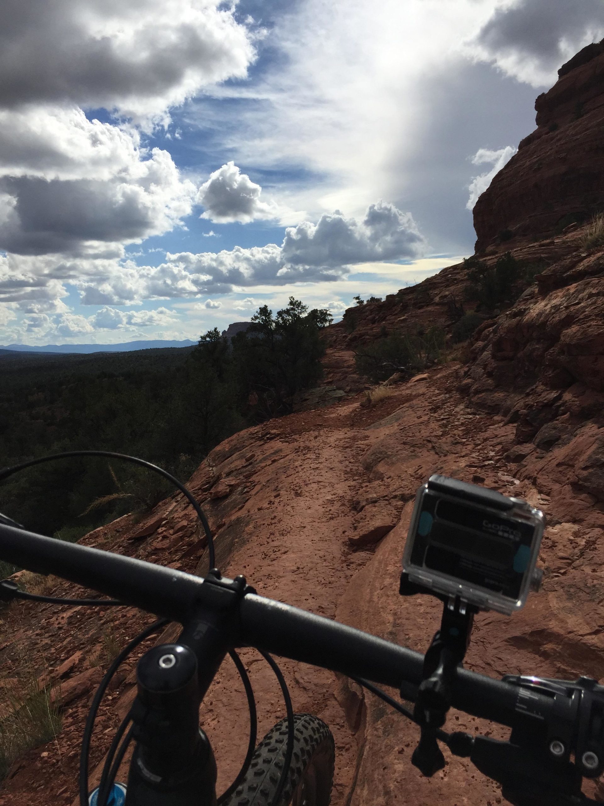 A close-up view of a mountain bike handlebar, with a GoPro camera mounted on it, set against a backdrop of a rocky trail. The sky is filled with fluffy clouds, and the terrain features earthy tones of red rock and greenery, suggesting a remote outdoor biking adventure. Upper Dry Creek Area Trails mountain bike trail.
