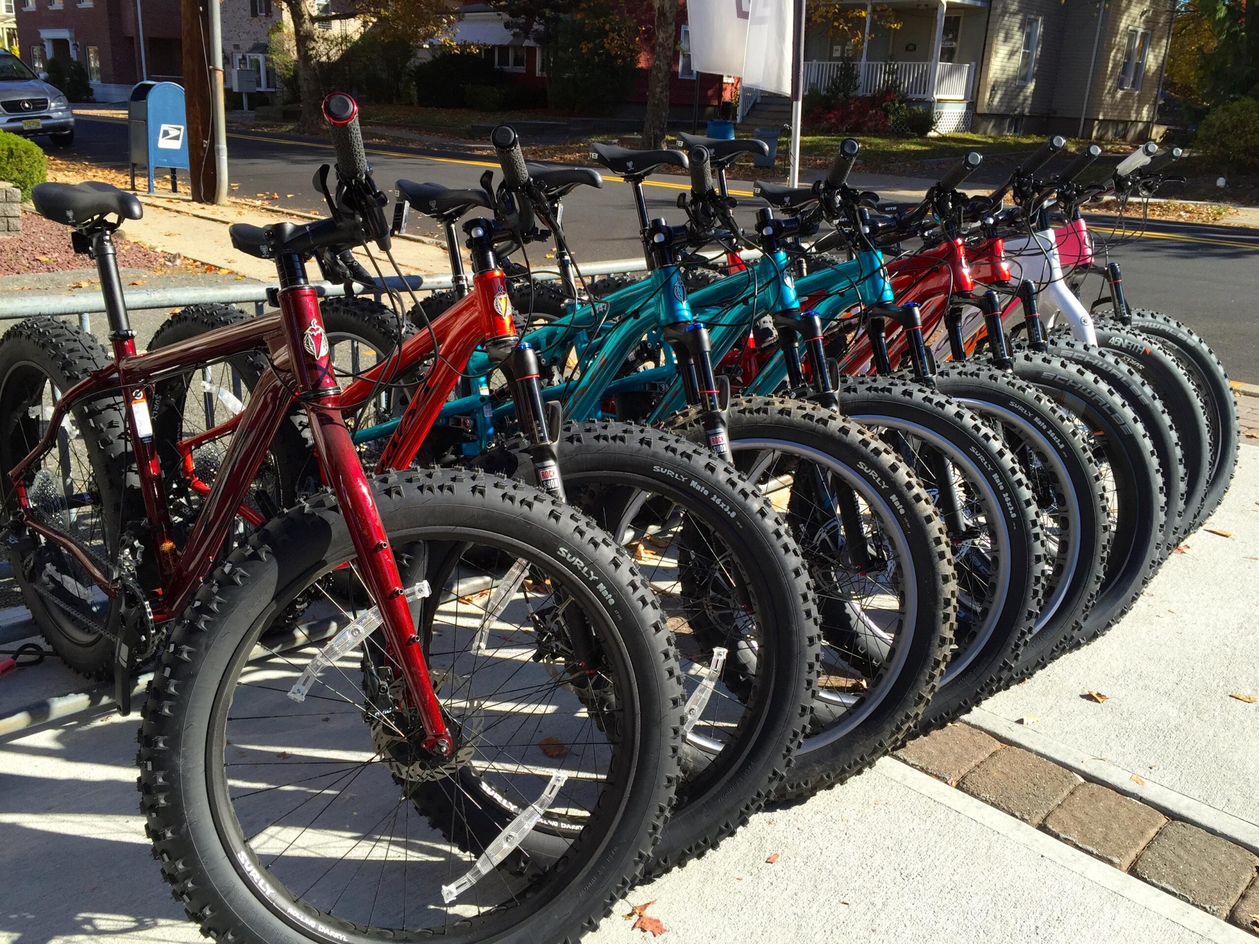 A row of colorful mountain bikes parked on a bike rack, showcasing a variety of frame colors including red, teal, and white. Each bike features wide, textured tires designed for off-road riding. The background includes a mailbox and residential buildings, indicating an outdoor setting.