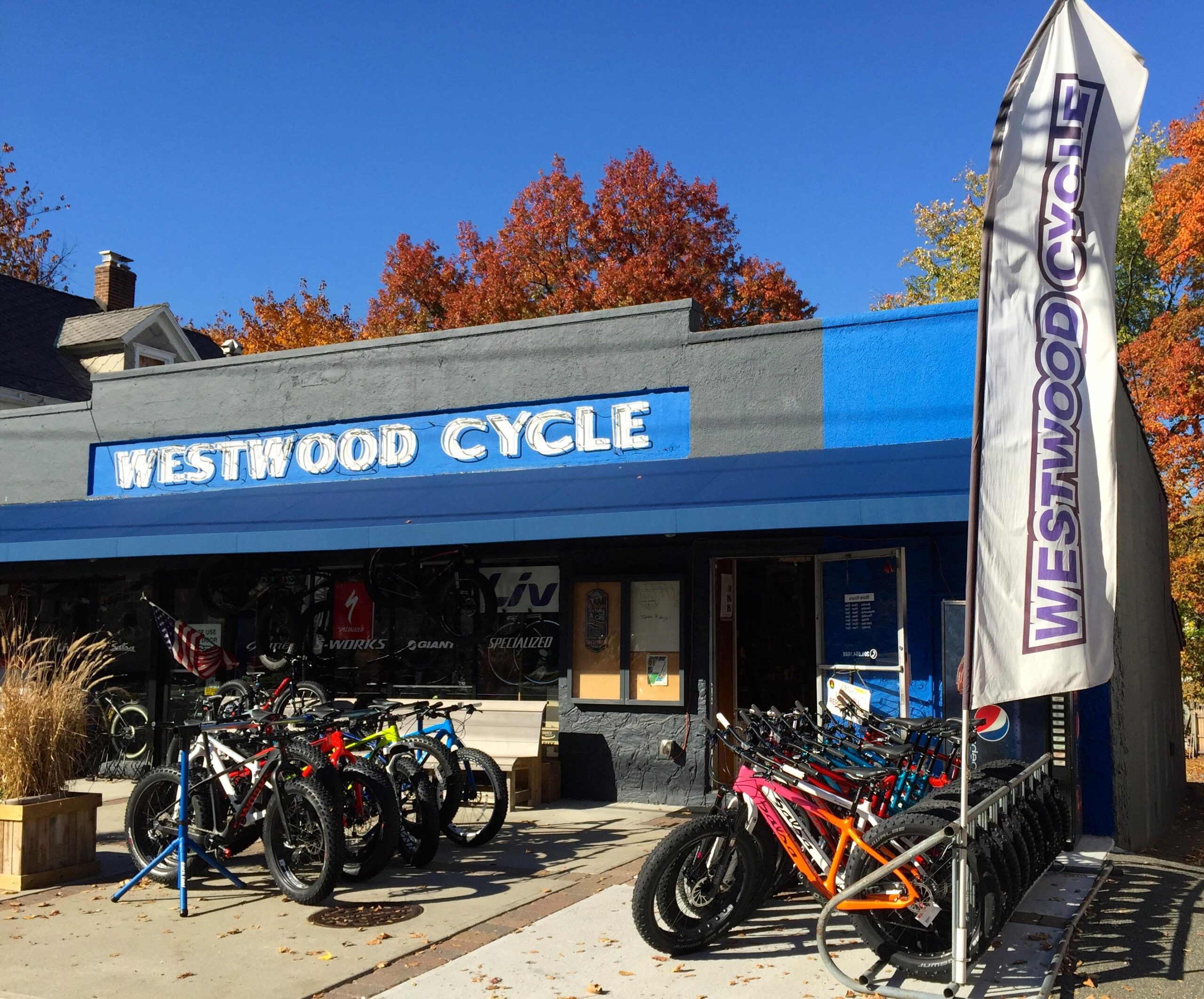 Alt text: "Exterior of Westwood Cycle bike shop featuring a blue facade, the store name prominently displayed, and several bicycles on display outside. Autumn trees with colorful foliage are visible in the background."