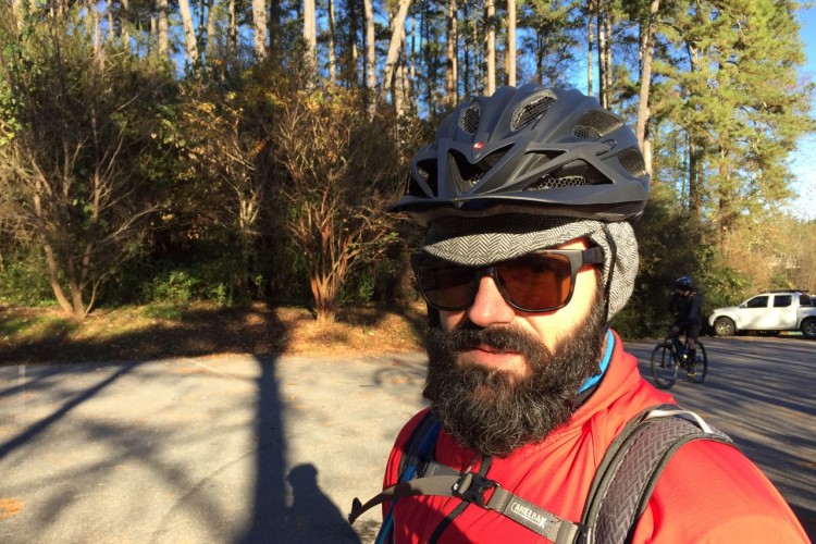 A man with a thick beard wearing a cycling helmet and sunglasses stands in an outdoor setting, surrounded by trees. He is dressed in a red cycling jacket and appears to be ready for a bike ride. In the background, another cyclist can be seen on a trail. The scene is sunny and showcases a natural, wooded area.