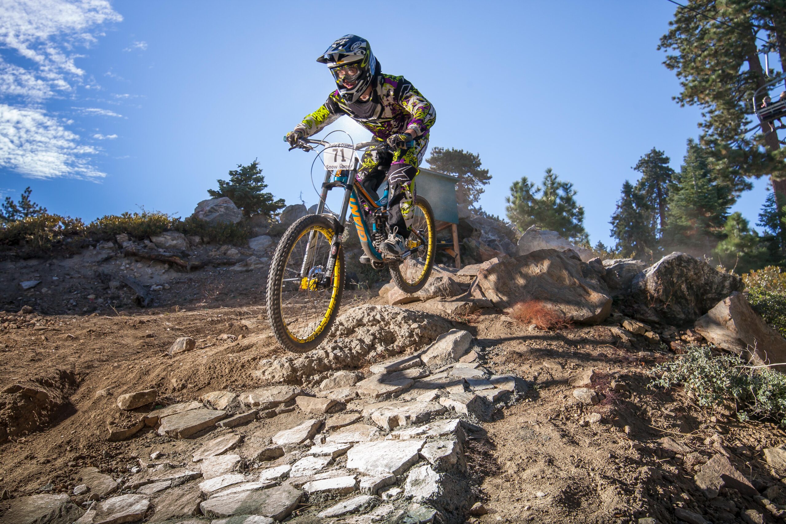 A mountain biker in protective gear jumps over a rocky section on a bike trail, with a blue sky and scattered clouds in the background. The terrain is rugged, featuring dirt and stone, surrounded by trees. Big Bear Mountain Resort mountain bike trail.