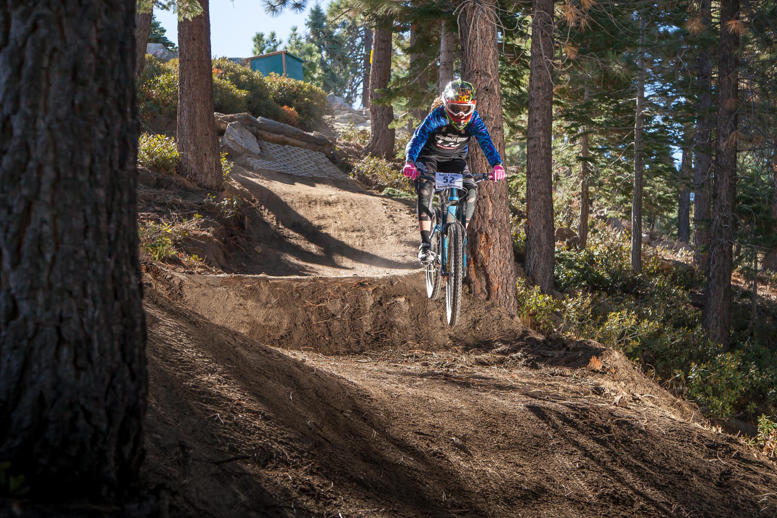 A mountain biker in colorful gear jumps off a dirt ramp on a forested trail, surrounded by trees and underbrush. The rider wears a helmet with a visor and is mid-air, showcasing skillful maneuvering. Big Bear Mountain Resort mountain bike trail.