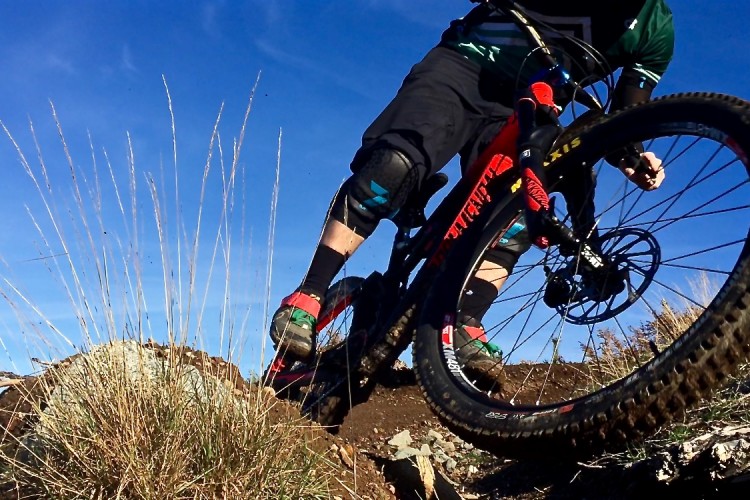 A mountain biker in protective gear maneuvers around a rocky trail, showcasing dynamic action against a clear blue sky. The foreground features grass and dirt, emphasizing the rugged terrain.