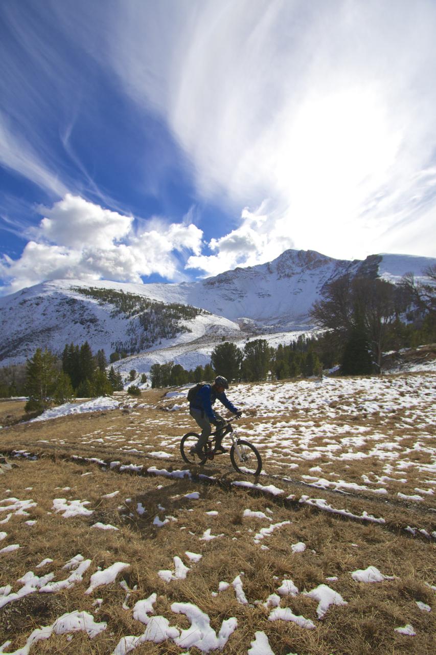 A person riding a mountain bike on a snowy trail in a mountainous landscape, with green trees and patches of snow on the ground. The sky is blue with white, wispy clouds. Rocky Creek Loop mountain bike trail.