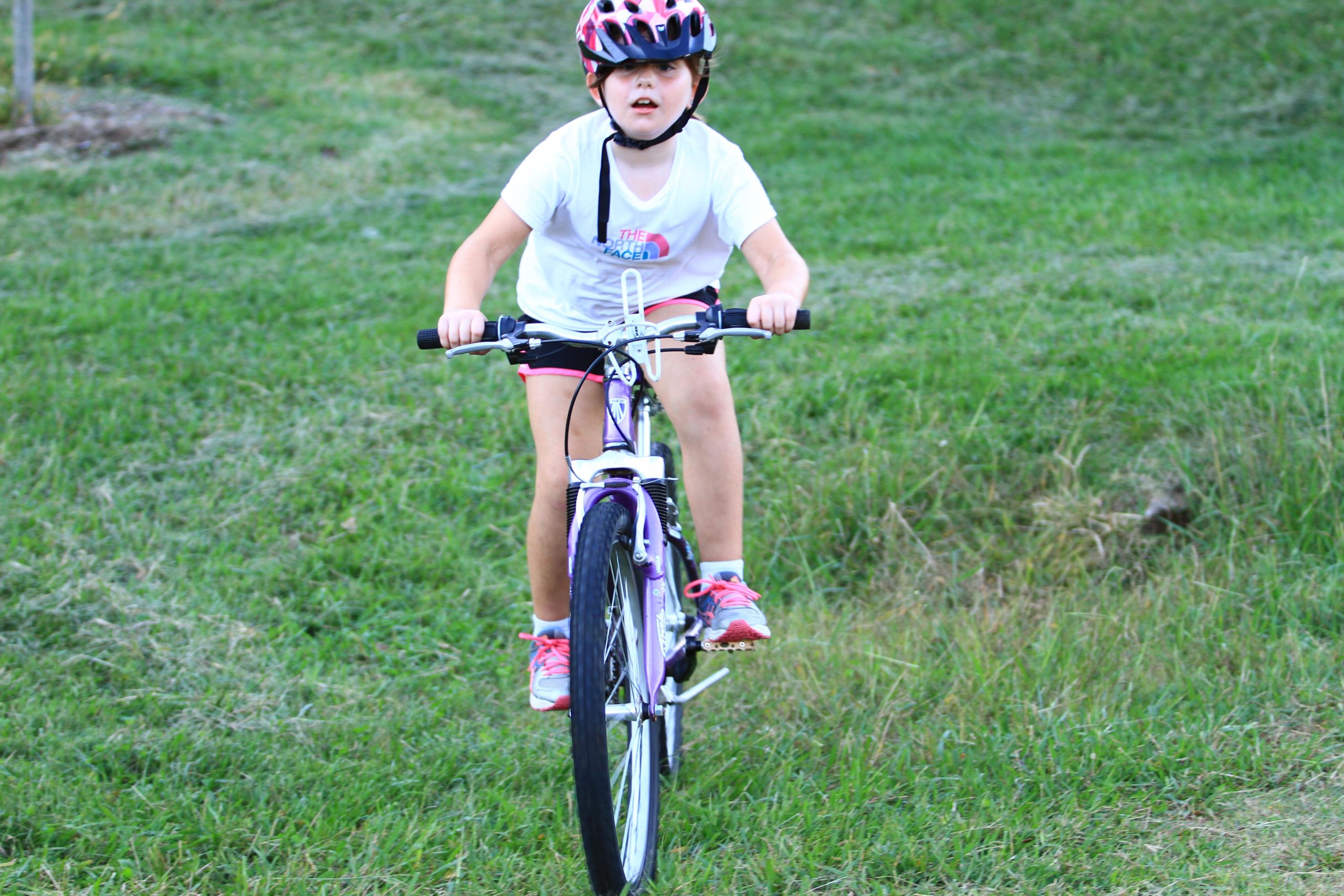 A young child riding a bicycle on green grass, wearing a pink helmet, a white t-shirt, and black shorts. The child appears focused and determined while pedaling. Tanglewood Park mountain bike trail.