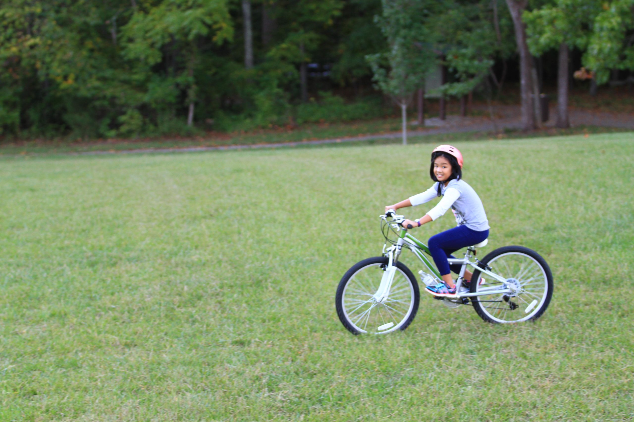 A young girl riding a bicycle across a grassy field, smiling and wearing a pink helmet. In the background, trees provide a natural setting. Tanglewood Park mountain bike trail.