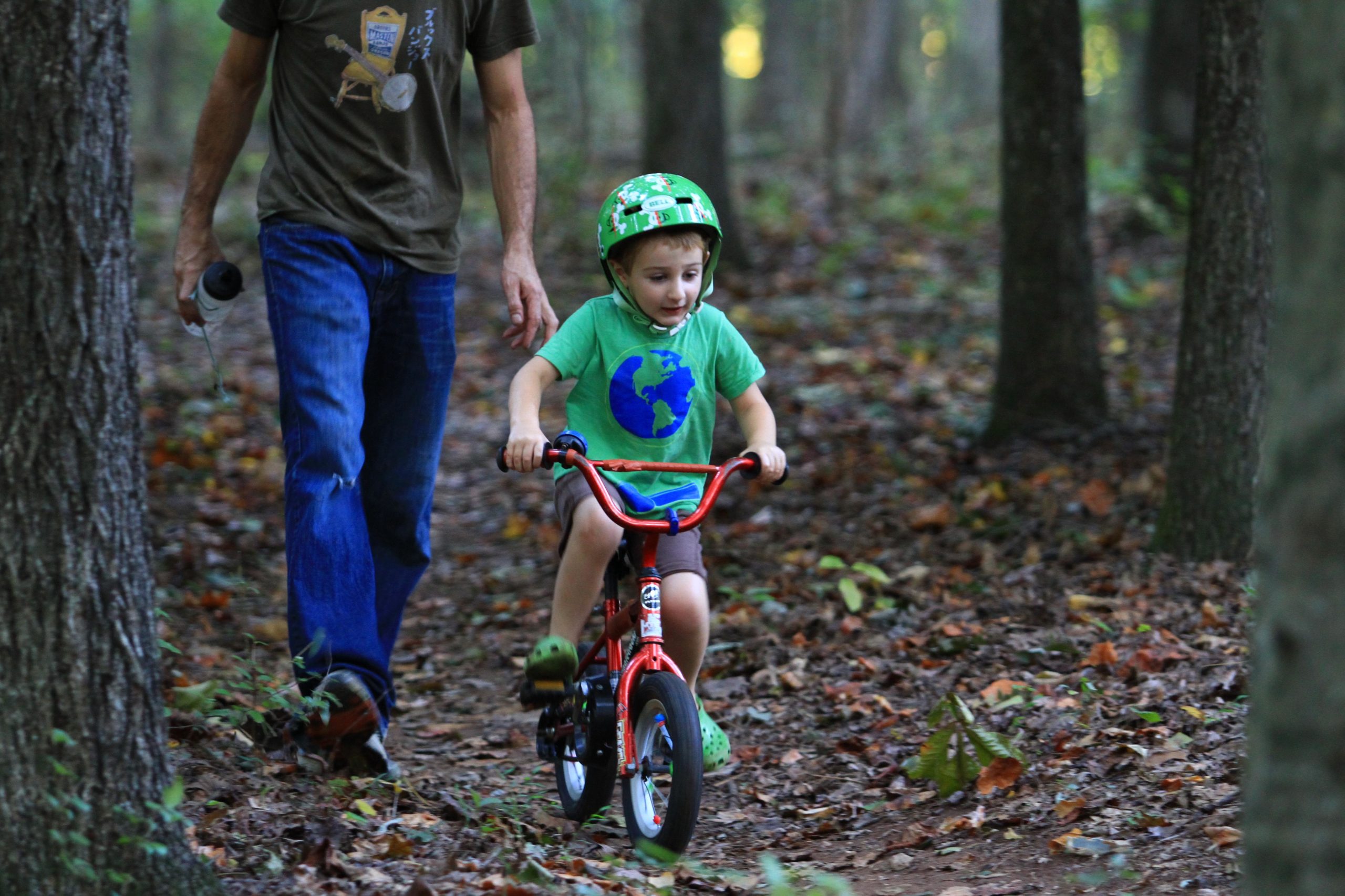A young child riding a small red bicycle on a dirt path in a wooded area, while an adult walks alongside, holding a drink. The child wears a green helmet and a shirt featuring a globe design, displaying a joyful expression as they navigate the bike. The scene is surrounded by trees and fallen leaves, illustrating an outdoor setting. Tanglewood Park mountain bike trail.