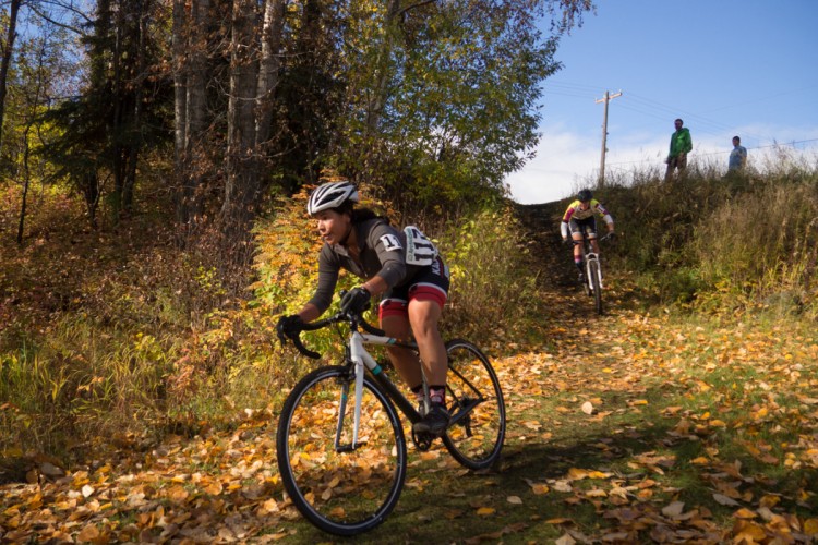A cyclist rides along a foliage-covered trail in an autumn environment, with colorful leaves scattered on the ground. Another cyclist descends a hill in the background, while two spectators watch from the side. The scene takes place on a sunny day with blue skies and a hint of greenery in the surrounding trees.