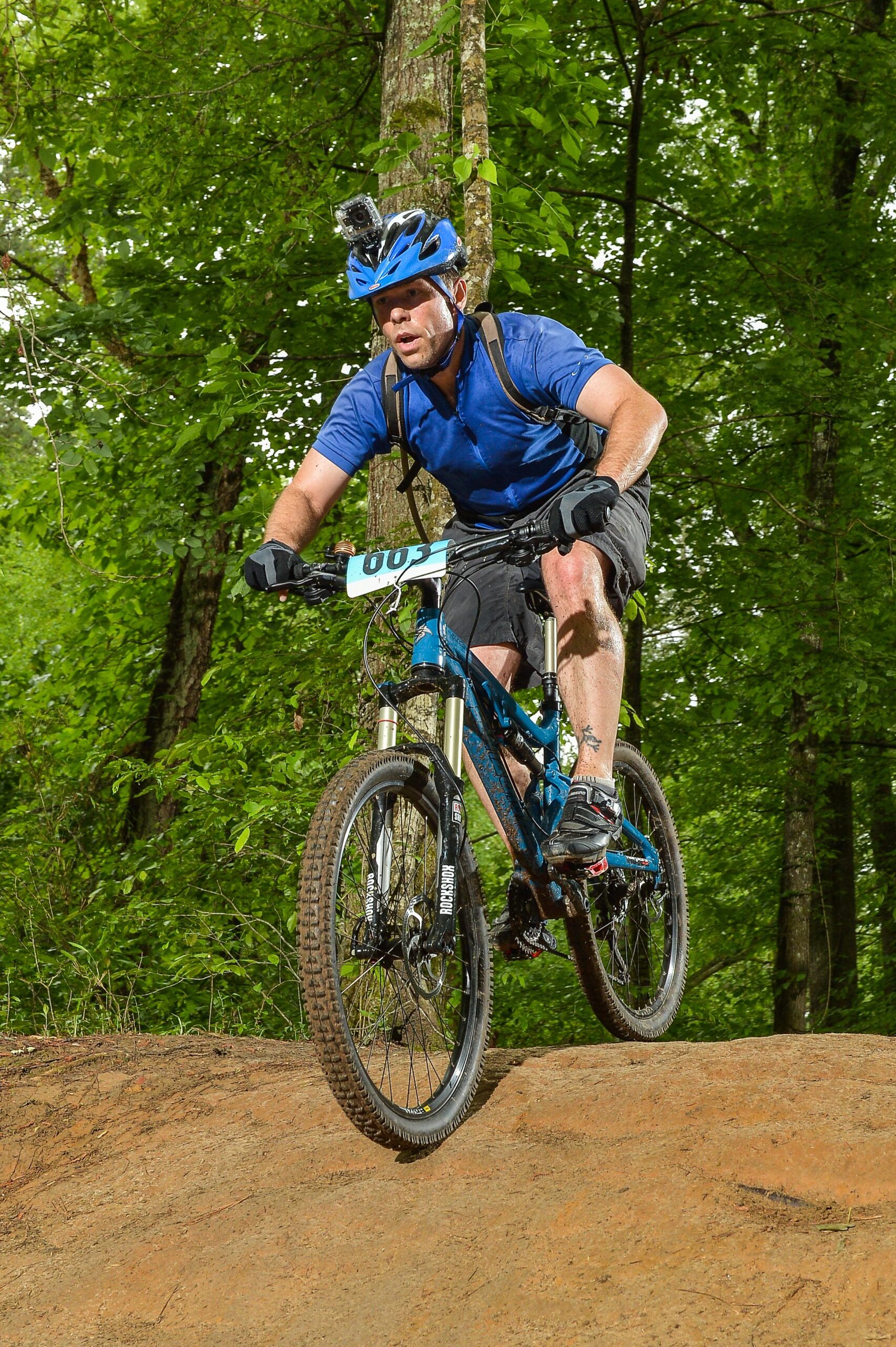 Santa Cruz Heckler: A mountain biker wearing a blue shirt, black shorts, and a blue helmet with a camera on top rides down a dirt slope in a forested area. The biker looks focused and determined as they navigate the terrain surrounded by green foliage and trees. The bike has a race number attached to it.
