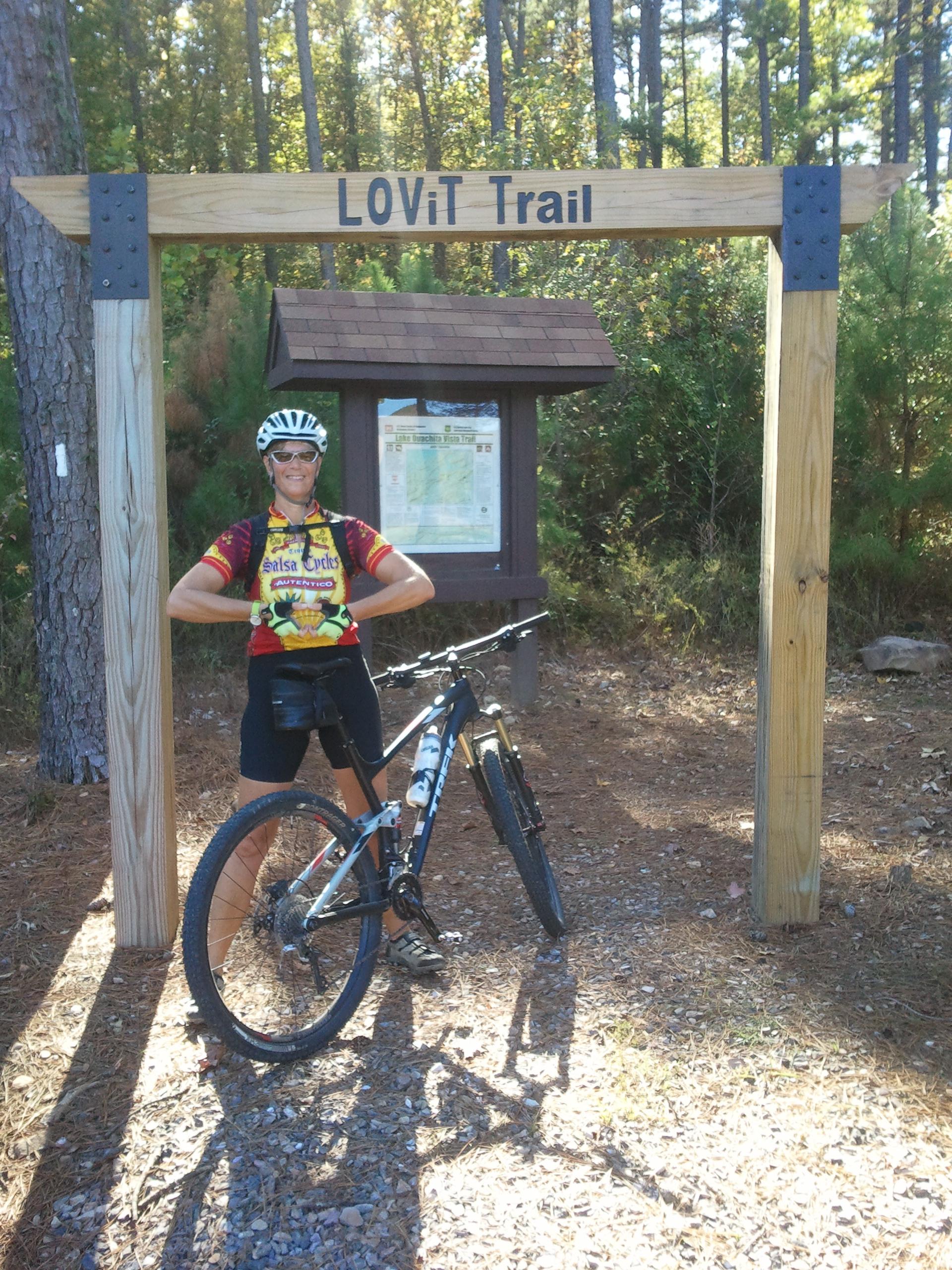 A cyclist in a colorful jersey poses confidently in front of the LOViT Trail entrance. They are standing beside a mountain bike, with a trail map visible on the sign behind them, surrounded by trees and natural scenery. Lake Ouachita Vista Trail (LOViT) mountain bike trail.