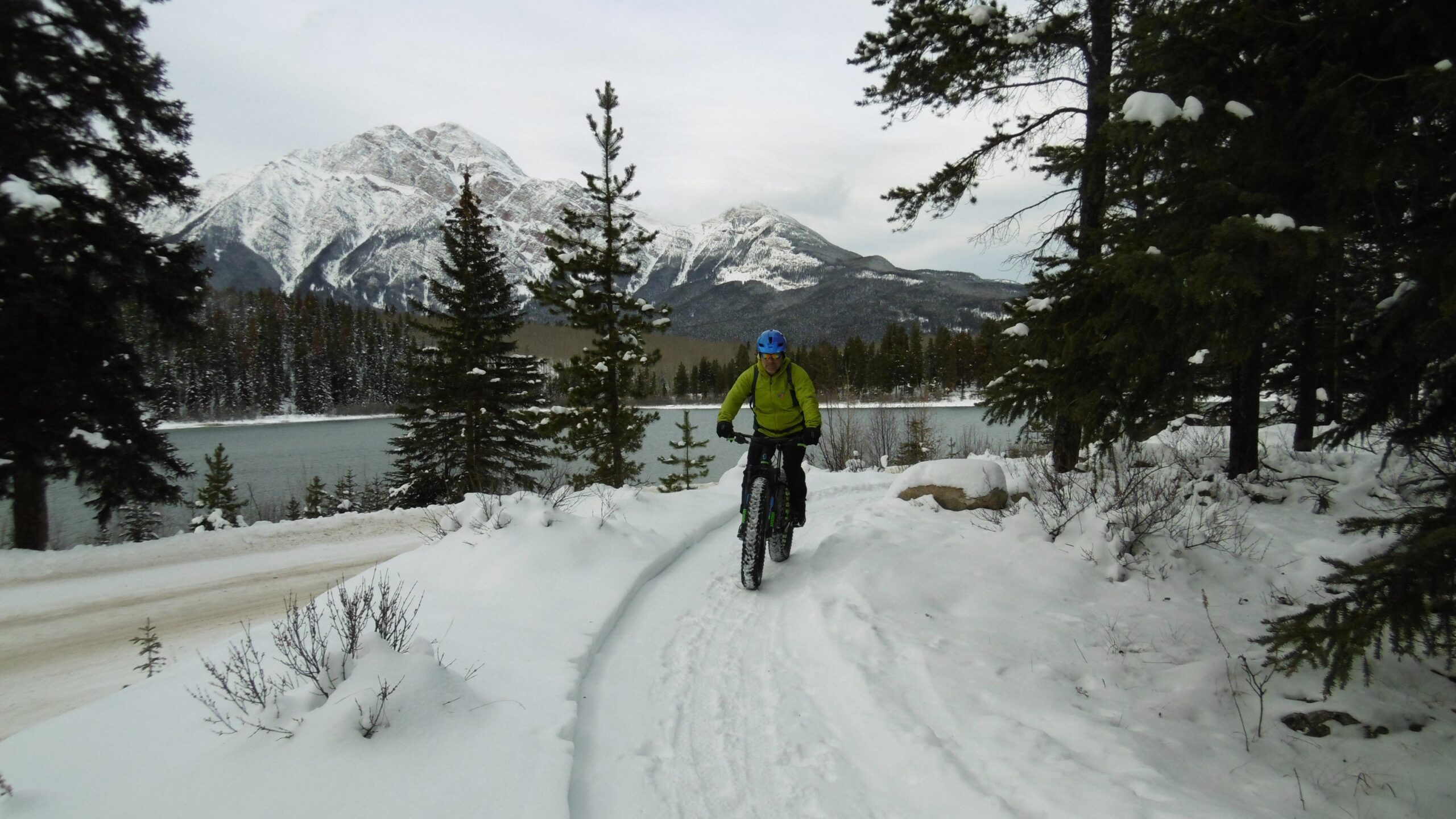 A person wearing a bright green jacket and blue helmet rides a fat bike along a snowy path near a lake, with snow-covered mountains and evergreen trees in the background. The scene captures a winter landscape with a tranquil atmosphere. #15 - Catholic Church to Pyramid Lake return mountain bike trail.
