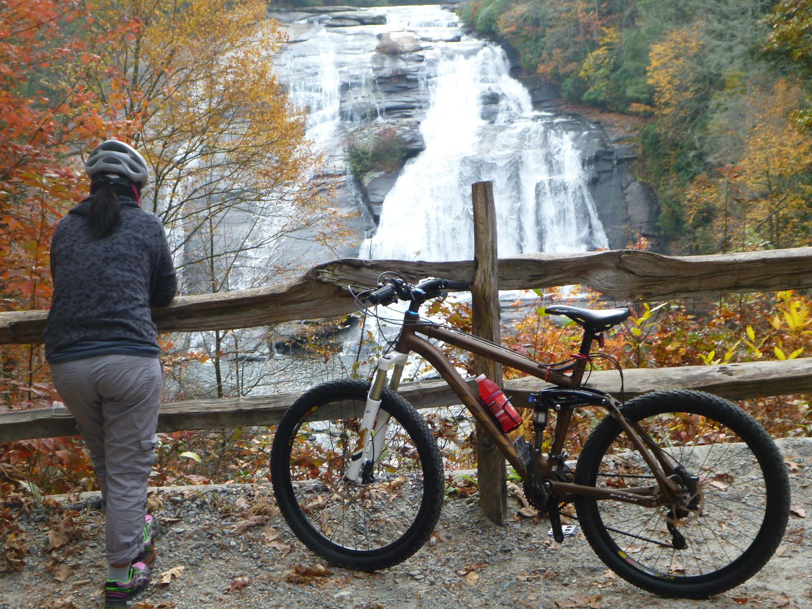 A person in a gray sweater and bike helmet leans against a wooden fence, gazing at a waterfall surrounded by autumn foliage. A mountain bike is parked beside them on a gravel path. DuPont State Forest mountain bike trail.