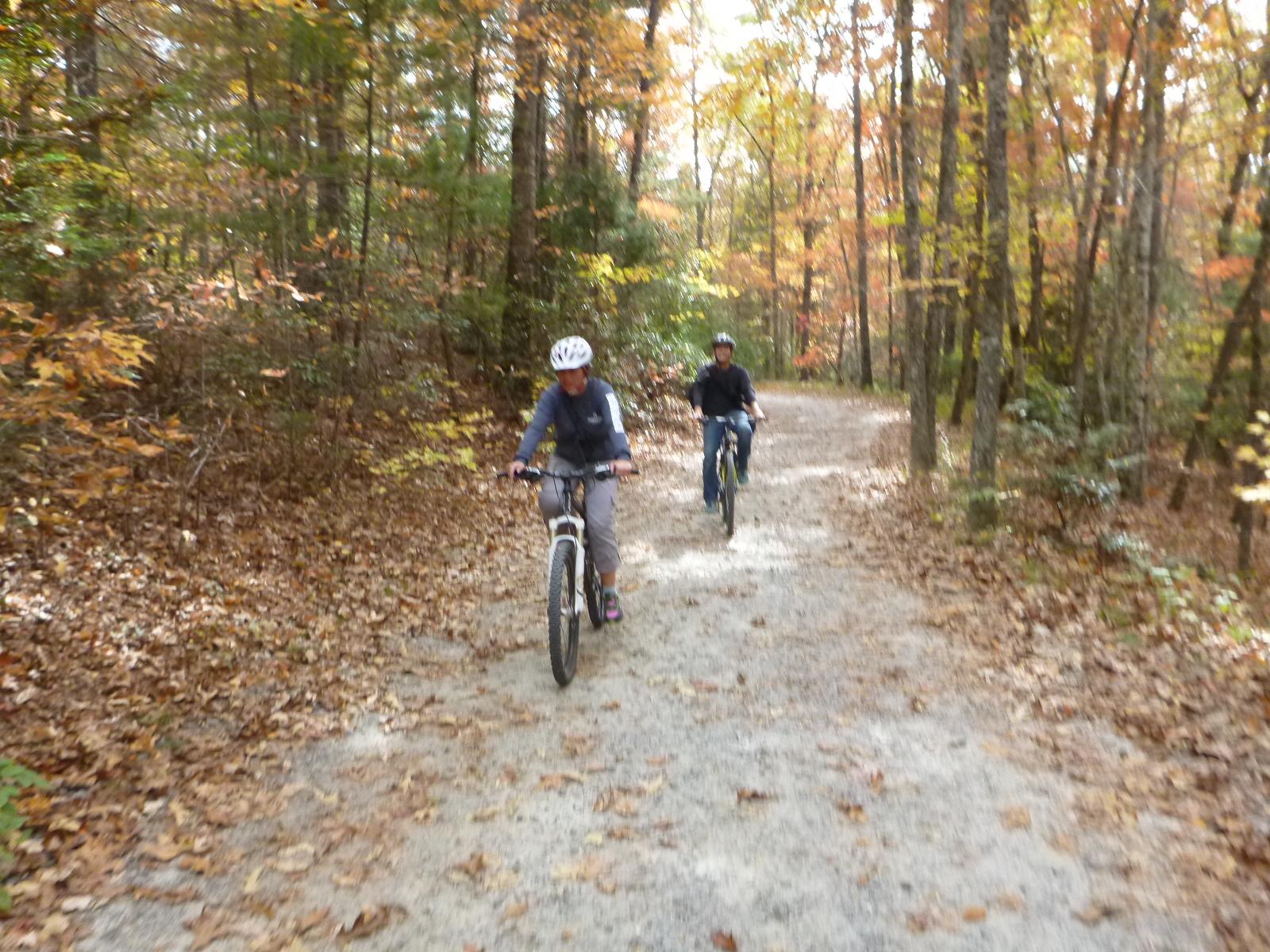 Two people riding bicycles along a gravel path surrounded by trees in autumn, with colorful leaves scattered on the ground and on the trees. DuPont State Recreational Forest mountain bike trail.