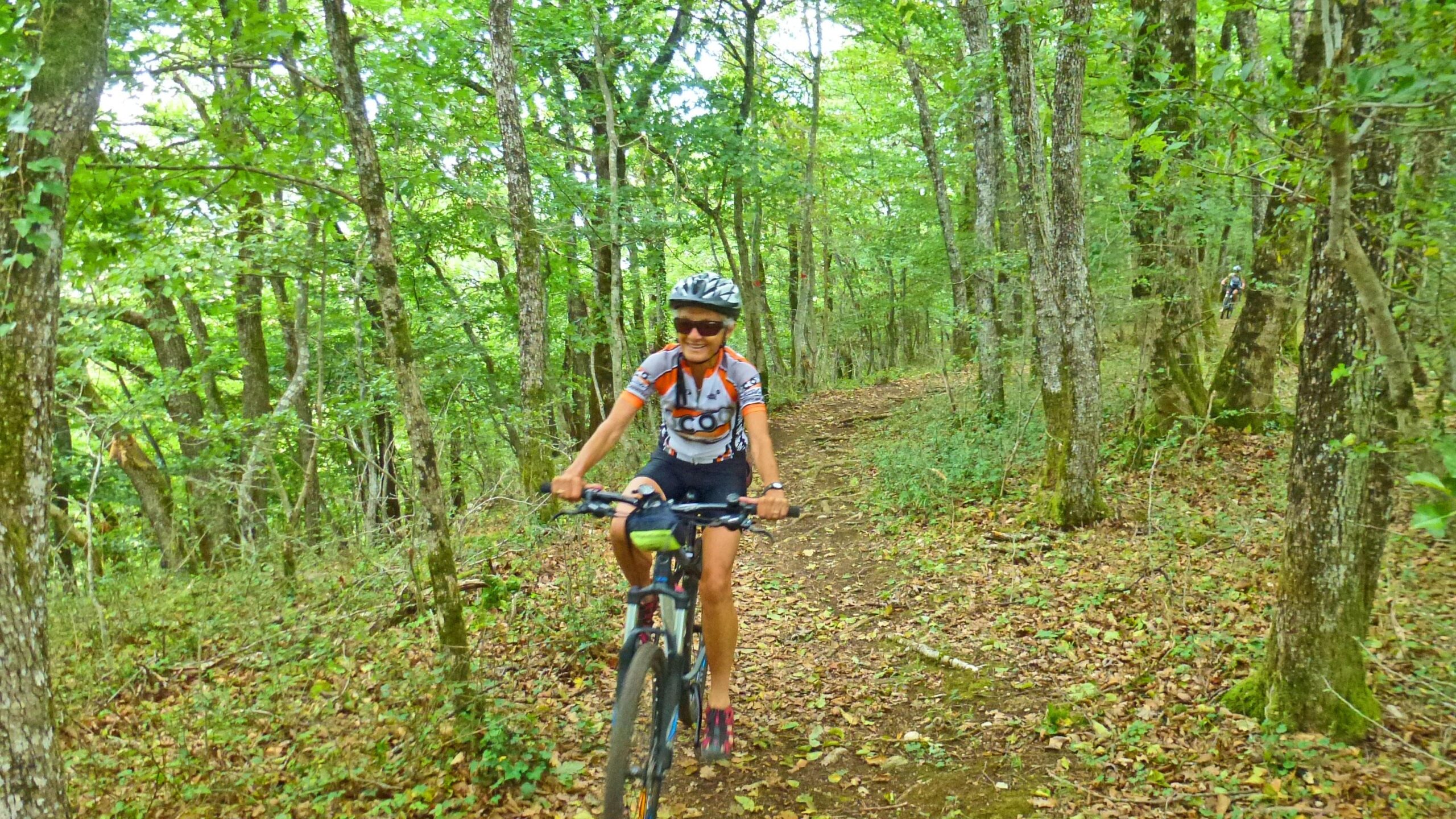 A person riding a mountain bike on a forest trail surrounded by green trees and fallen leaves, wearing a helmet and sunglasses, and sporting a cycling jersey. Great Jura Crossing mountain bike trail.