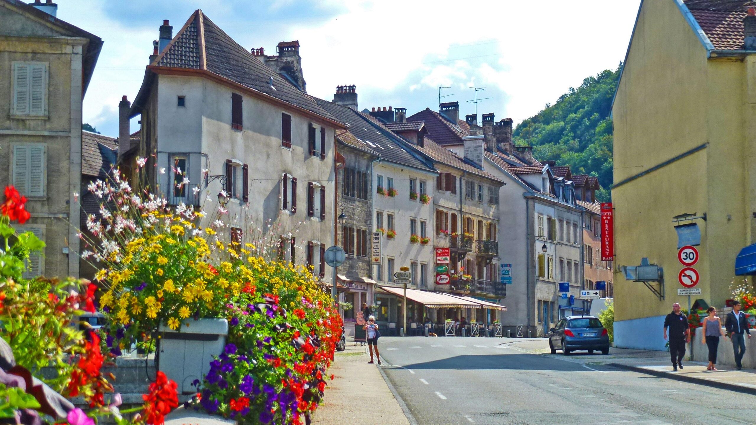 A picturesque village street featuring charming old buildings with flower boxes, vibrant flowerbeds in the foreground, and a few pedestrians strolling along the sidewalk. A car is parked nearby, and there are signs visible, hinting at local businesses. The scene is set against a backdrop of rolling hills under a partly cloudy sky. Great Jura Crossing mountain bike trail.