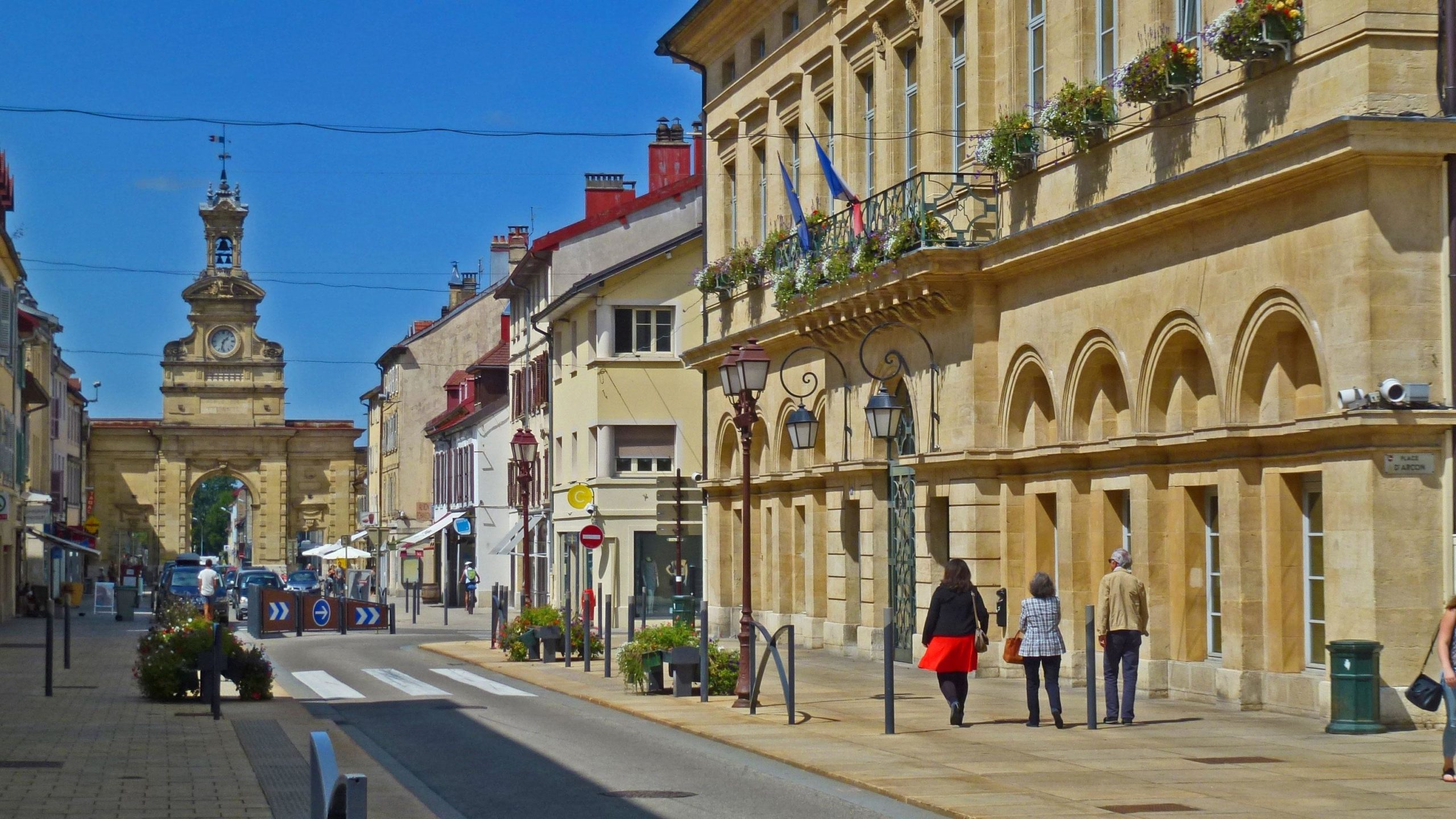 A picturesque street scene featuring historic buildings and a clock tower in the distance. The sun is shining brightly in a clear blue sky, and people are walking along the sidewalk. Flower boxes adorn the balconies of the buildings, while vintage street lamps line the street. A traffic circle with directional arrows is visible in the foreground. Great Jura Crossing mountain bike trail.
