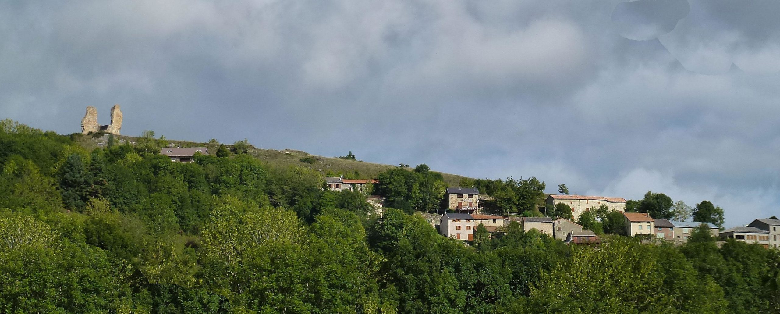 A panoramic view of a hilly landscape featuring dense green foliage and a small village. In the background, two stone towers rise on a hilltop under a partly cloudy sky, while several houses with varied roofs are scattered across the lower slope. Great Crossing of the Ariege mountain bike trail.