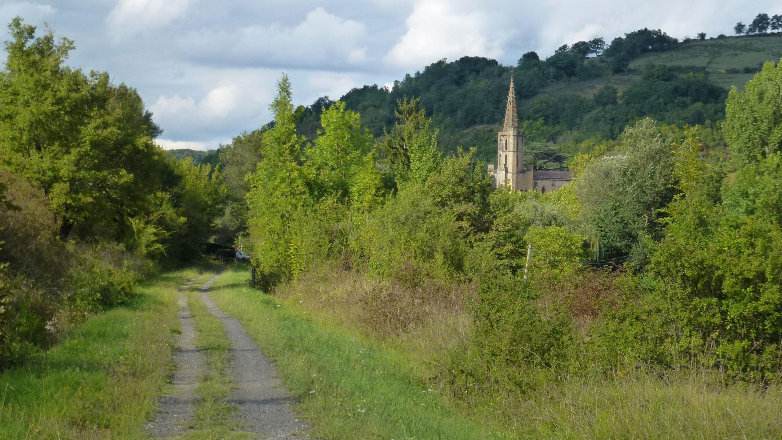 A serene pathway surrounded by lush greenery, leading towards a distant church steeple visible among the trees, set against a backdrop of rolling hills and partly cloudy skies. Great Crossing of the Ariege mountain bike trail.