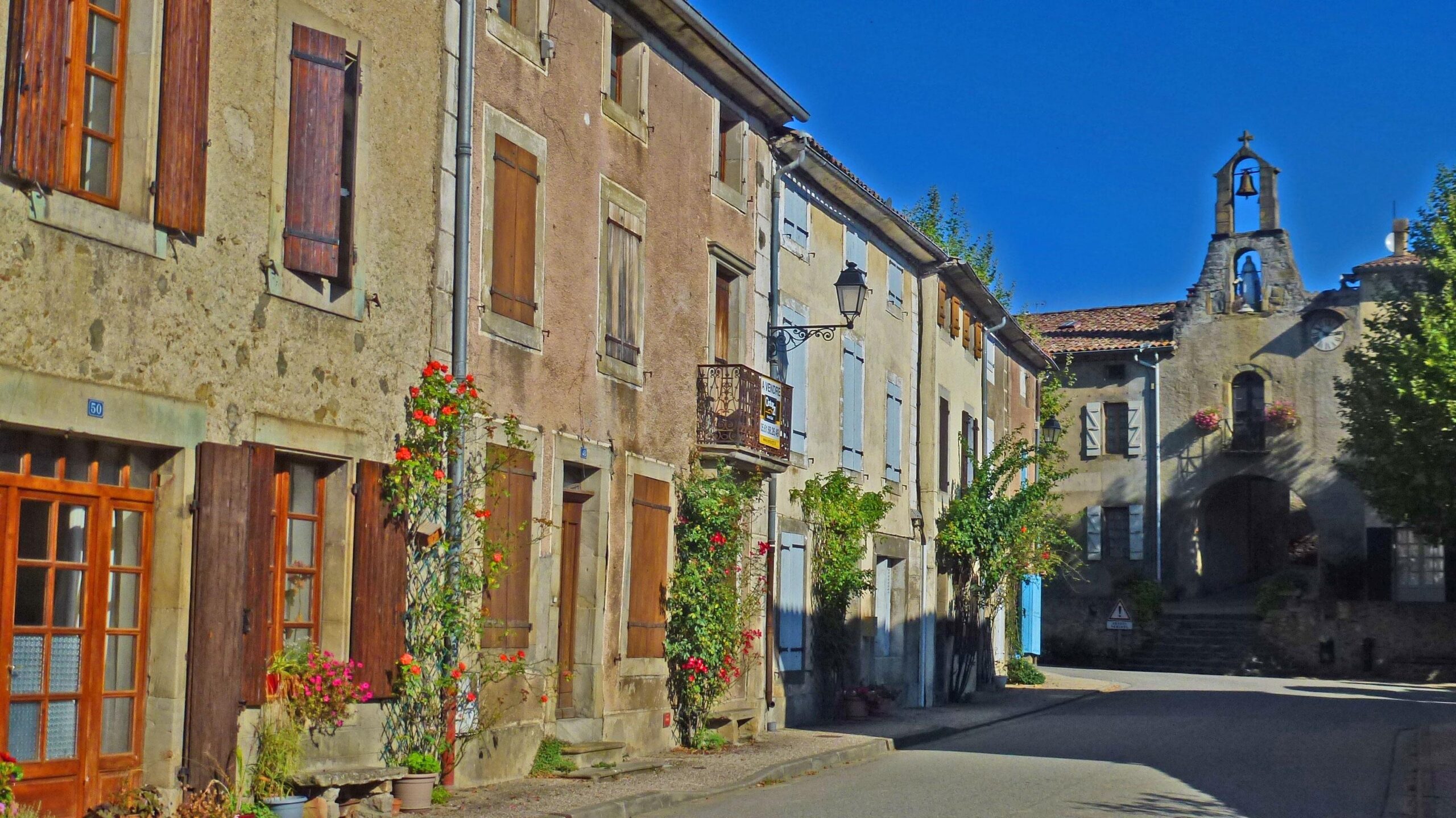A picturesque street scene featuring quaint, traditional stone buildings with wooden shutters and colorful flower pots lining the path. A charming church with a bell tower is visible at the end of the road, under a clear blue sky. Great Crossing of the Ariege mountain bike trail.