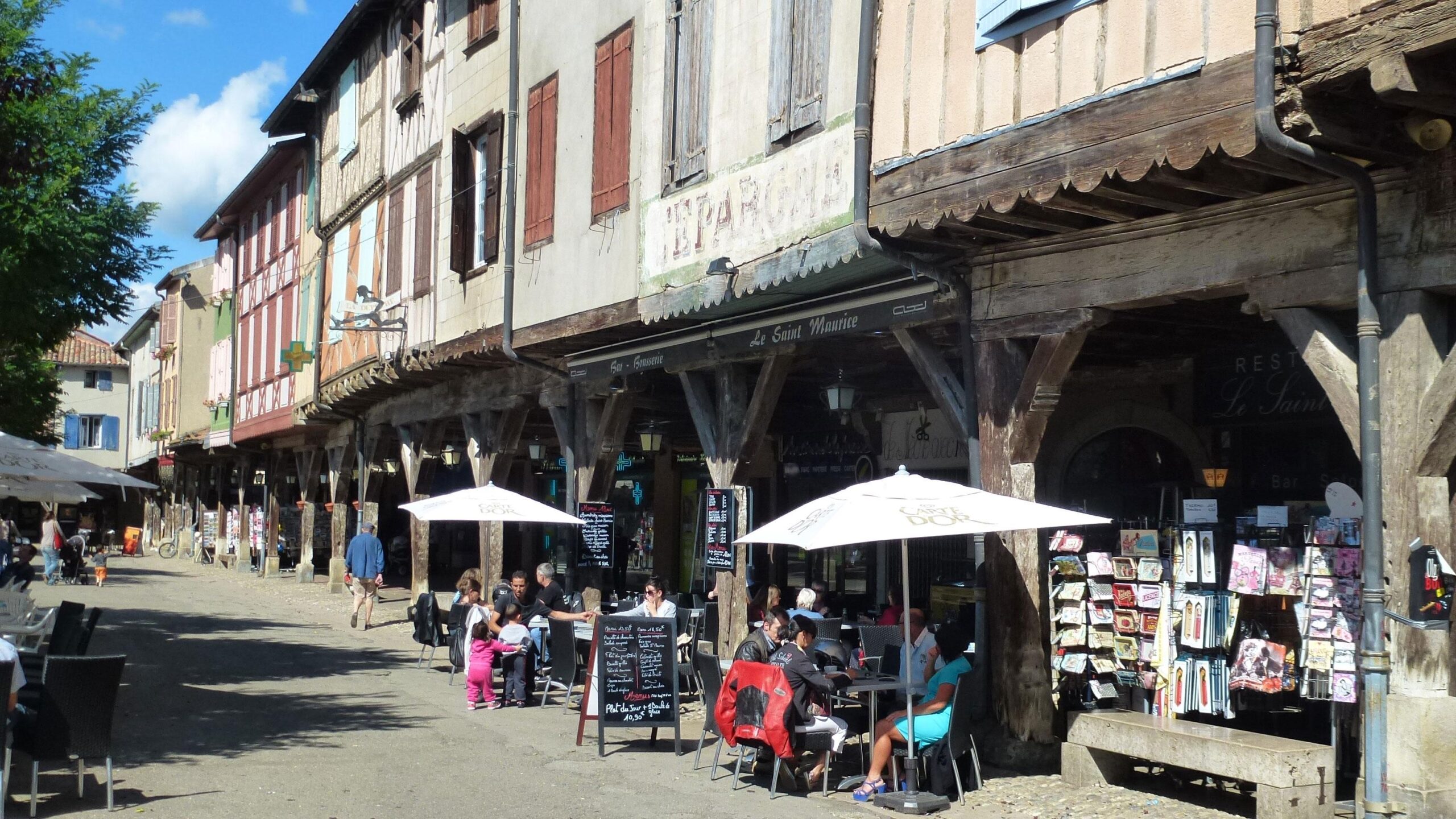 A picturesque street scene featuring traditional wooden-framed buildings with colorful shutters. In the foreground, people are seated at outdoor café tables under parasols, enjoying their meals. Nearby, a stand displays various items for sale. The bright blue sky and gentle cloud cover enhance the lively atmosphere of the area. Great Crossing of the Ariege mountain bike trail.