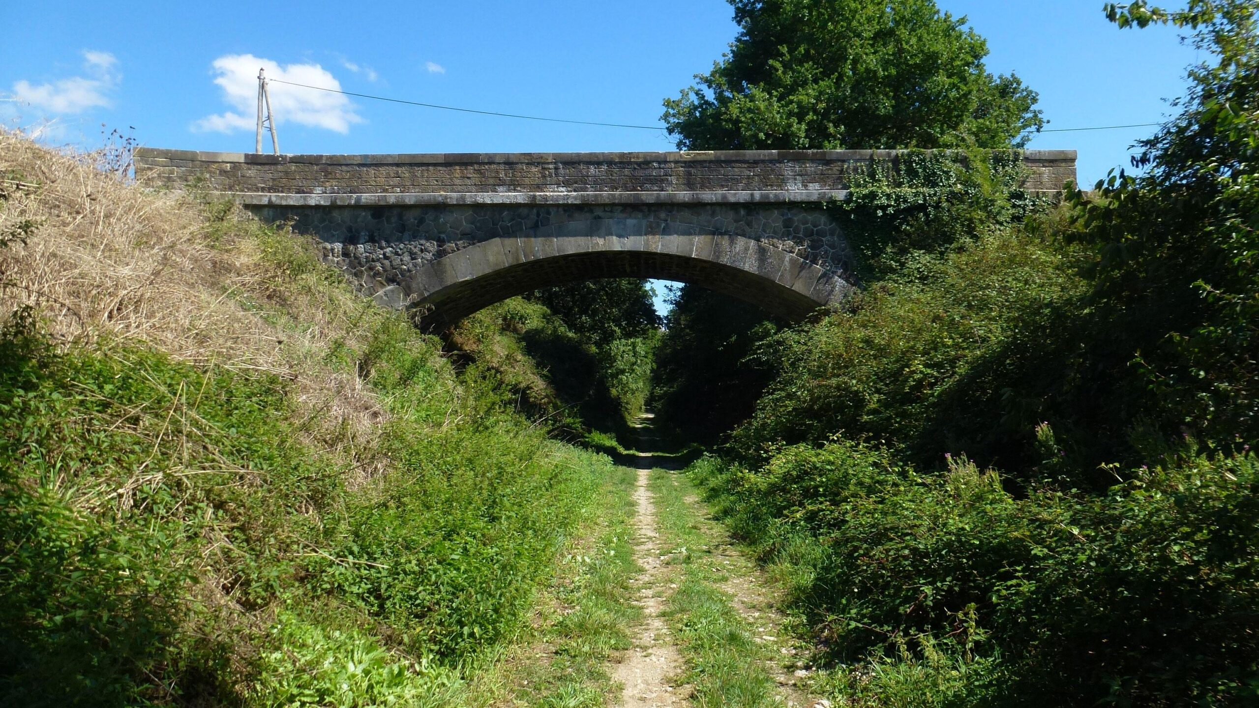 A stone bridge arching over a grassy path, surrounded by lush greenery and trees under a clear blue sky with a few clouds. The path leads through a natural corridor, inviting exploration. Great Crossing of the Ariege mountain bike trail.