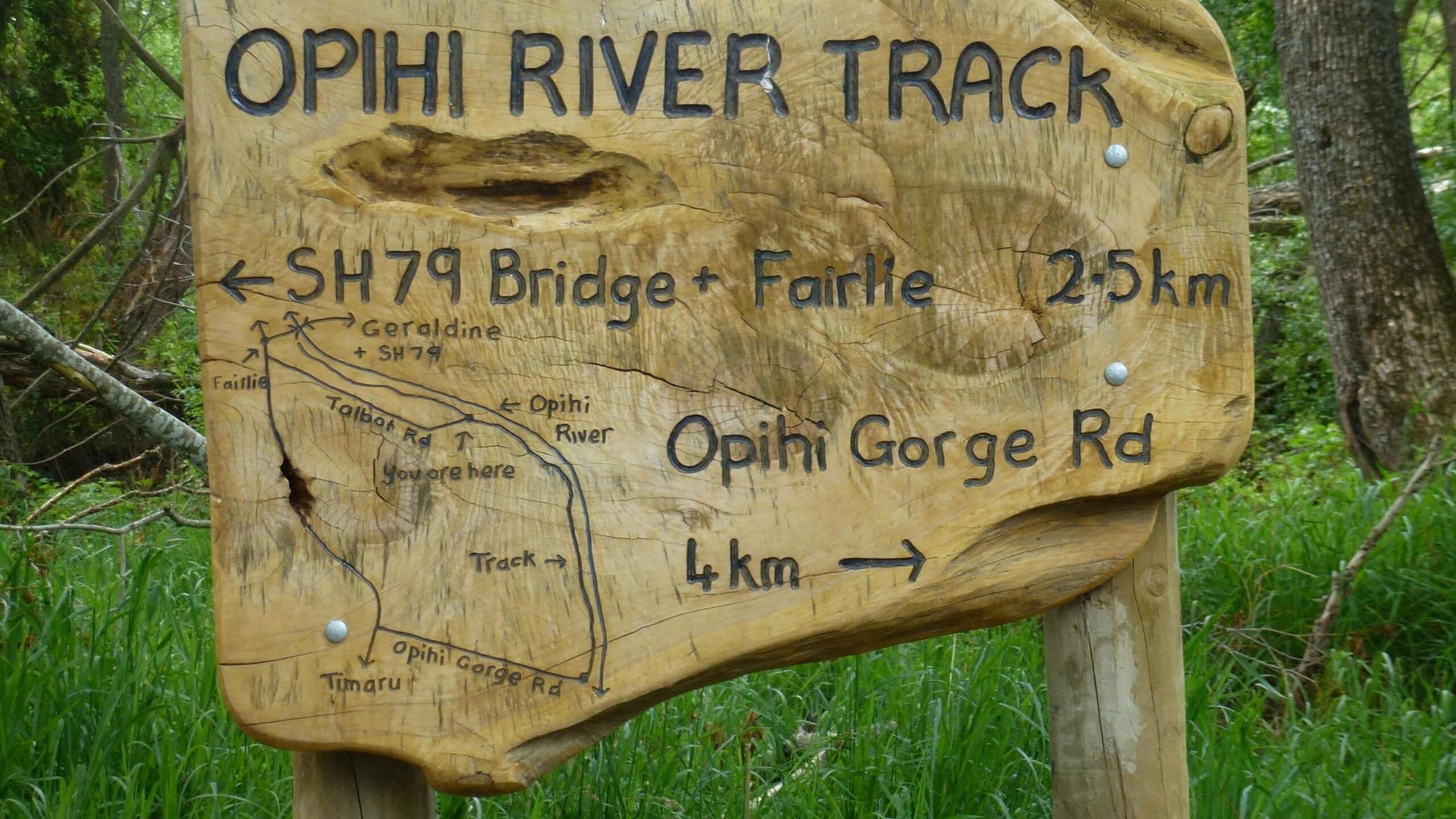Wooden signpost for the Opihi River Track, displaying directional arrows and distances to SH79 Bridge and Fairlie (2.5 km) and Opihi Gorge Road (4 km), with a map indicating the surrounding area and trails. Opihi River Trail mountain bike trail.