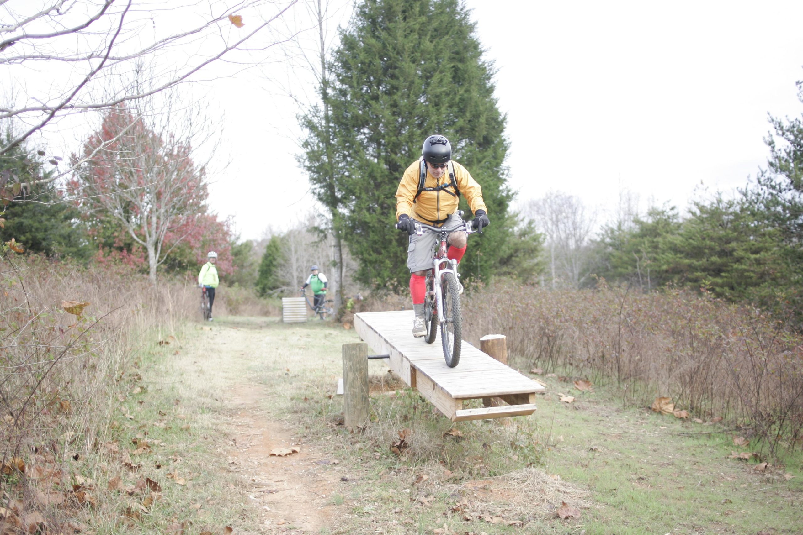 A mountain biker wearing a yellow jacket and helmet rides down a wooden ramp on a trail, with two other cyclists visible in the background. The scene is set in a natural landscape with trees and sparse foliage, indicating a cool, overcast day. Angler's Ridge mountain bike trail.