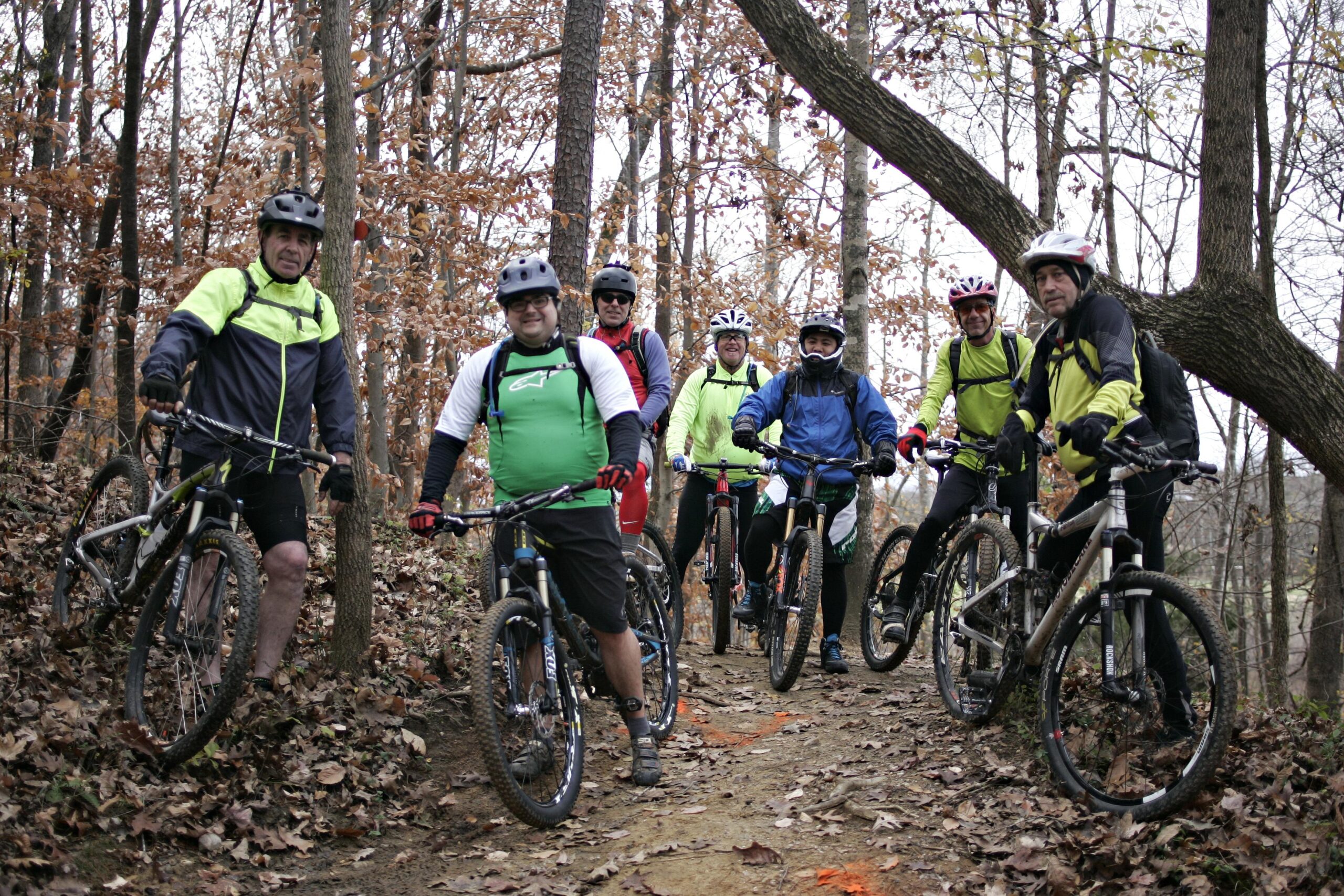 A group of eight mountain bikers posing on a wooded trail during autumn. They are wearing helmets and cycling gear, with their bikes parked beside them. The background features bare trees with some remaining autumn leaves on the ground. The atmosphere appears relaxed and friendly. Angler's Ridge mountain bike trail.