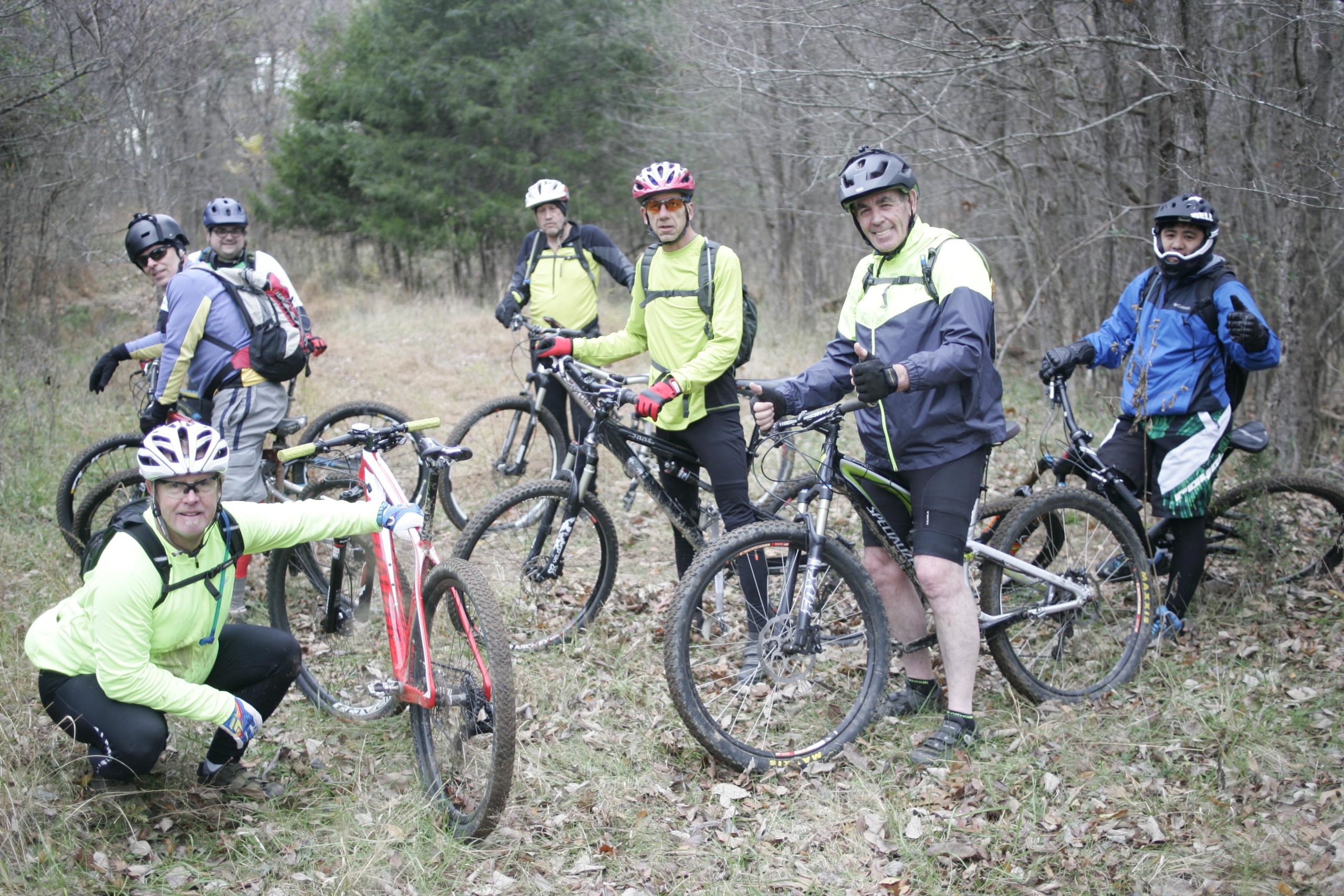 A group of seven mountain bikers dressed in bright jackets and helmets stands on a dirt trail surrounded by trees. One rider is crouched next to a red bike, while the others are posed around their bikes, smiling and giving thumbs up. The scene captures a moment of camaraderie and outdoor adventure. Angler's Ridge mountain bike trail.