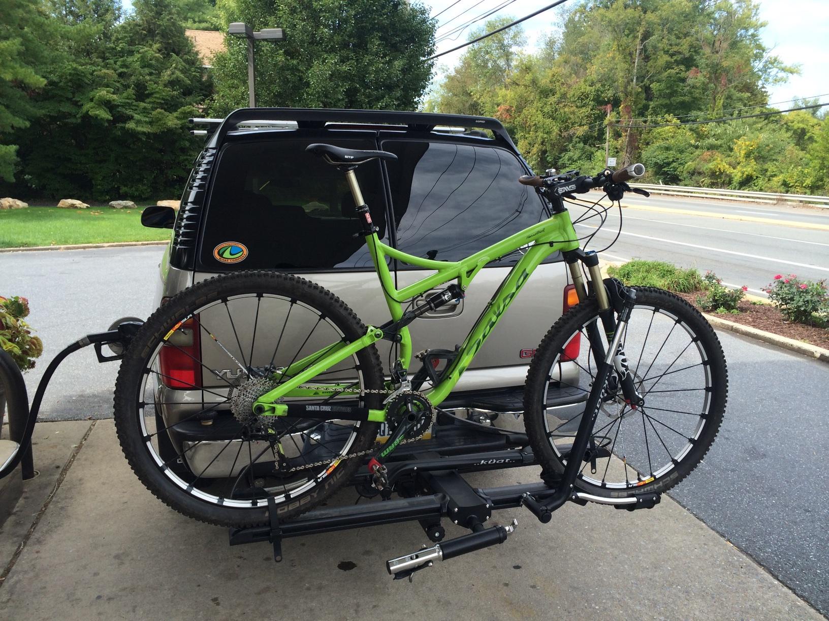 Salsa Horsethief: A bright green mountain bike secured to a hitch-mounted bike rack on the back of a silver vehicle. The background features a grassy area with shrubs and trees, and a clear sky overhead.