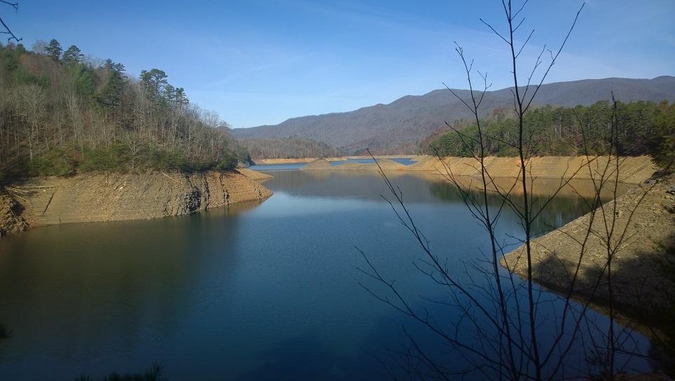 A serene landscape featuring a calm lake surrounded by steep, earthy banks and trees. The scene includes distant mountains under a clear blue sky, reflecting gently on the water's surface. Sparse vegetation with bare branches is visible in the foreground, adding to the natural beauty of the setting. Tsali Left Loop mountain bike trail.