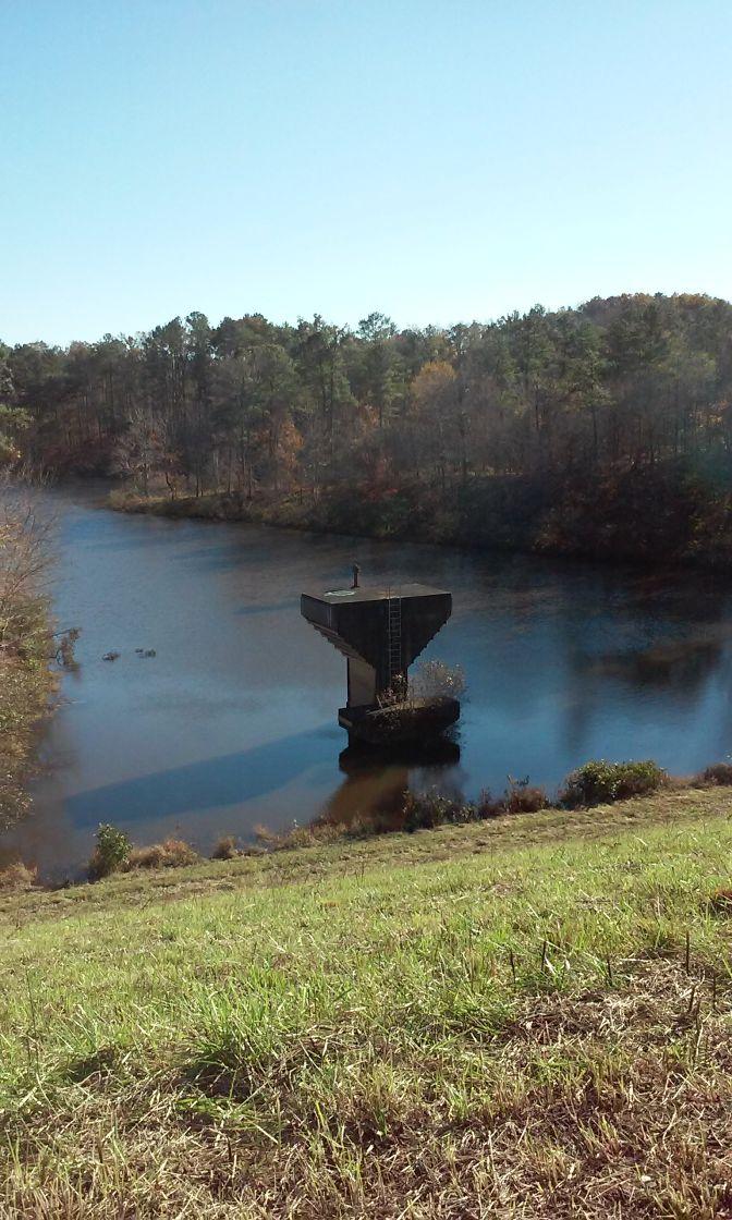 An abandoned concrete structure stands on a small island in a calm body of water, surrounded by trees displaying autumn colors. In the foreground, a grassy slope leads down to the water, where a person can be seen standing on the structure. The sky is clear and blue, creating a serene outdoor scene. Sylaward mountain bike trail.