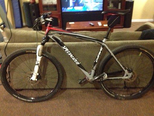 Specialized Carve Comp 29: A black and red mountain bike leaning against a couch, with a television in the background. The bike features thick tires and a lightweight frame, suitable for off-road cycling.