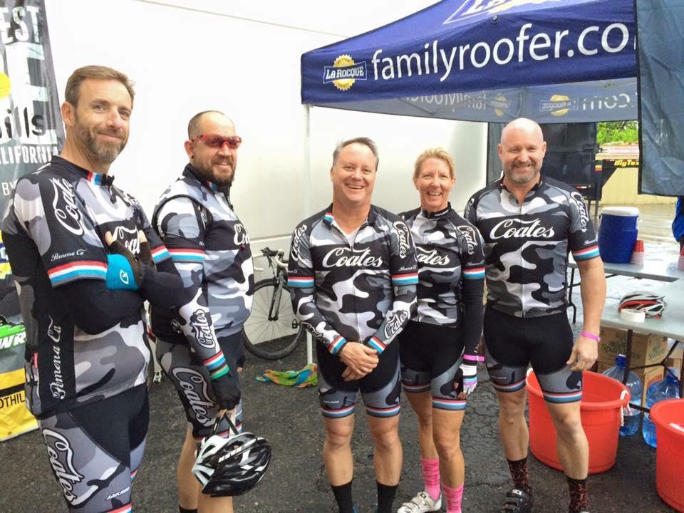 A group of five cyclists standing together, all wearing matching black and gray camouflage cycling jerseys with the name "Coates" on them. They are in a casual outdoor setting with a tent and bicycles in the background. The atmosphere appears friendly and spirited, likely at a cycling event or race.