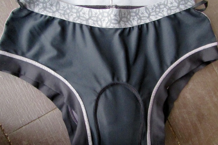 A pair of black women's underwear with a patterned gray waistband, featuring a textured fabric and a decorative edge along the legs. The underwear is laid flat on a wooden surface.