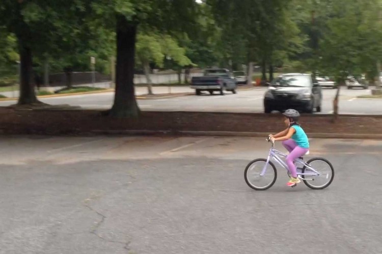 A young child wearing a helmet rides a purple bicycle in an empty parking lot, surrounded by trees and a few parked cars in the background. The child is observed mid-ride, showcasing a moment of joy and activity.