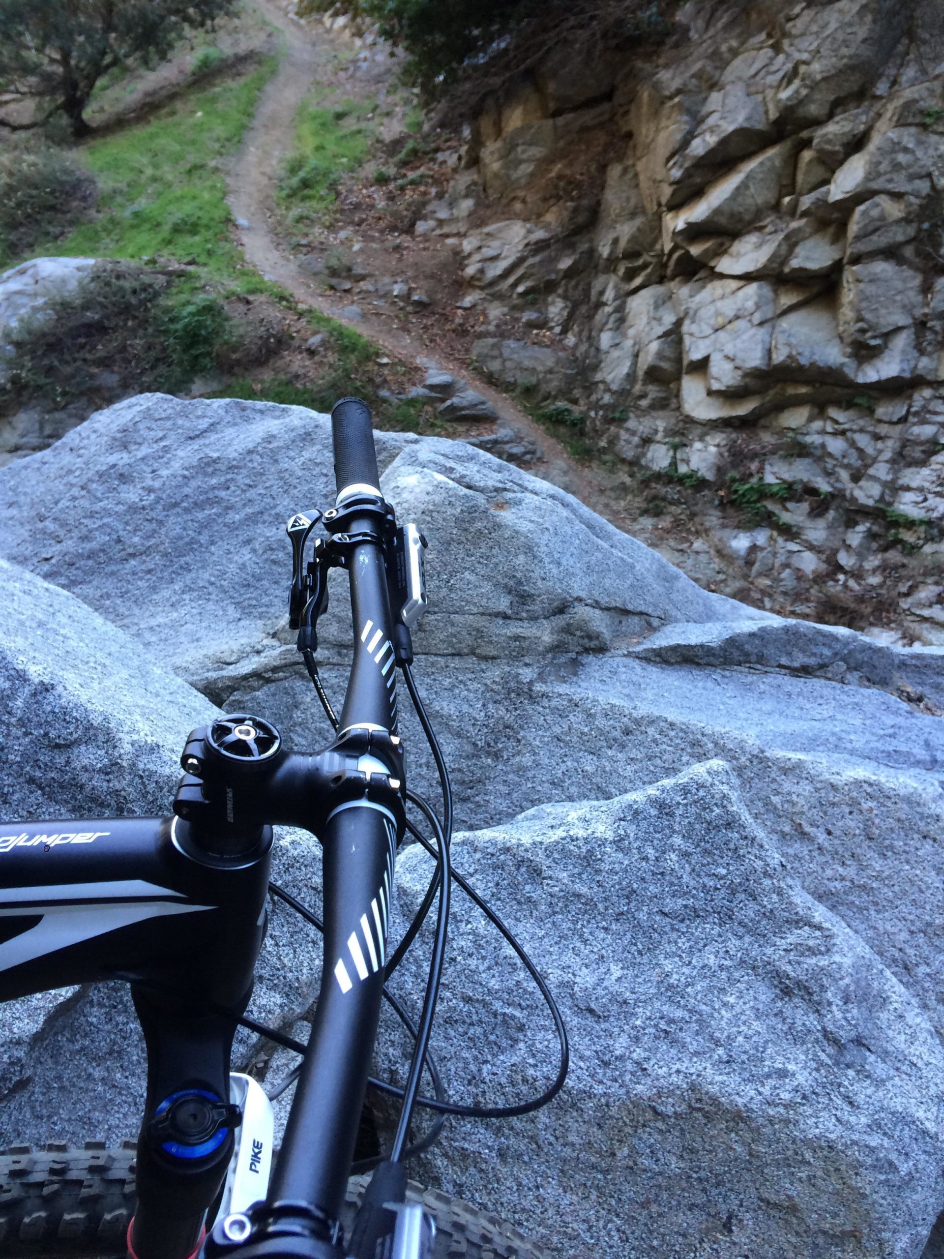 Close-up view of a mountain bike handlebar resting on large rocks, with a dirt trail winding through a green landscape in the background. The bike's components are visible, showcasing the handlebars and stem against the rugged terrain. Mount Wilson Trail mountain bike trail.