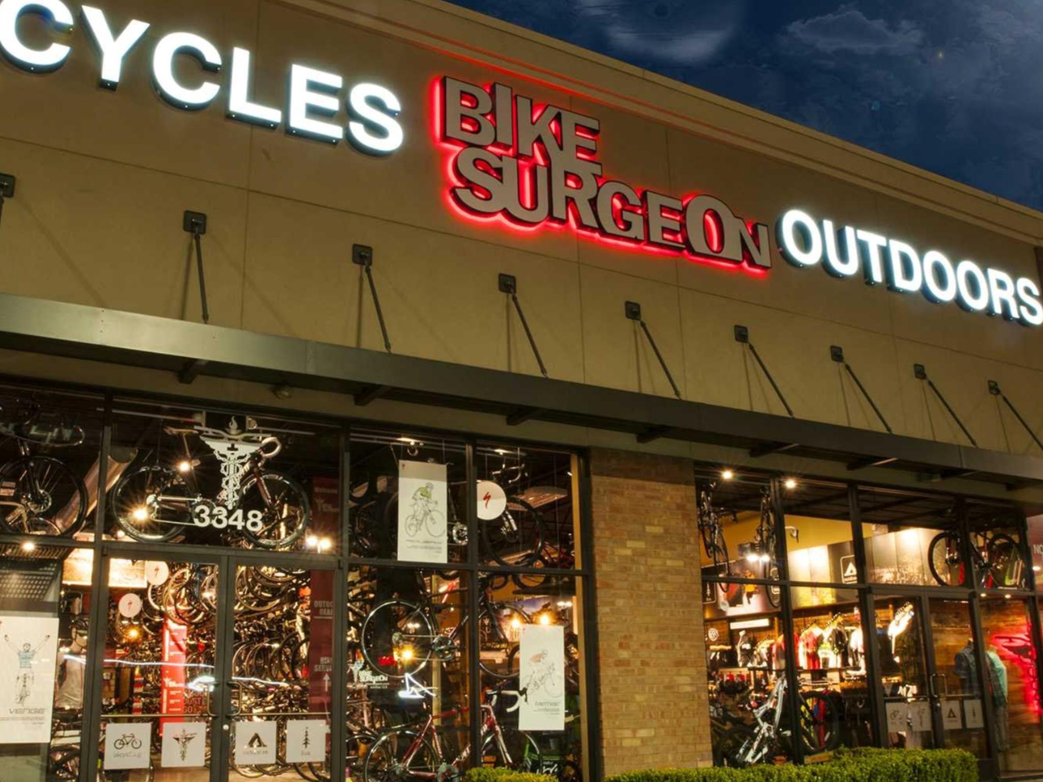 Exterior view of a bike shop named "Bike Surgeon Outdoors," featuring bright signage and a window display showcasing various bicycles and cycling gear. The shop