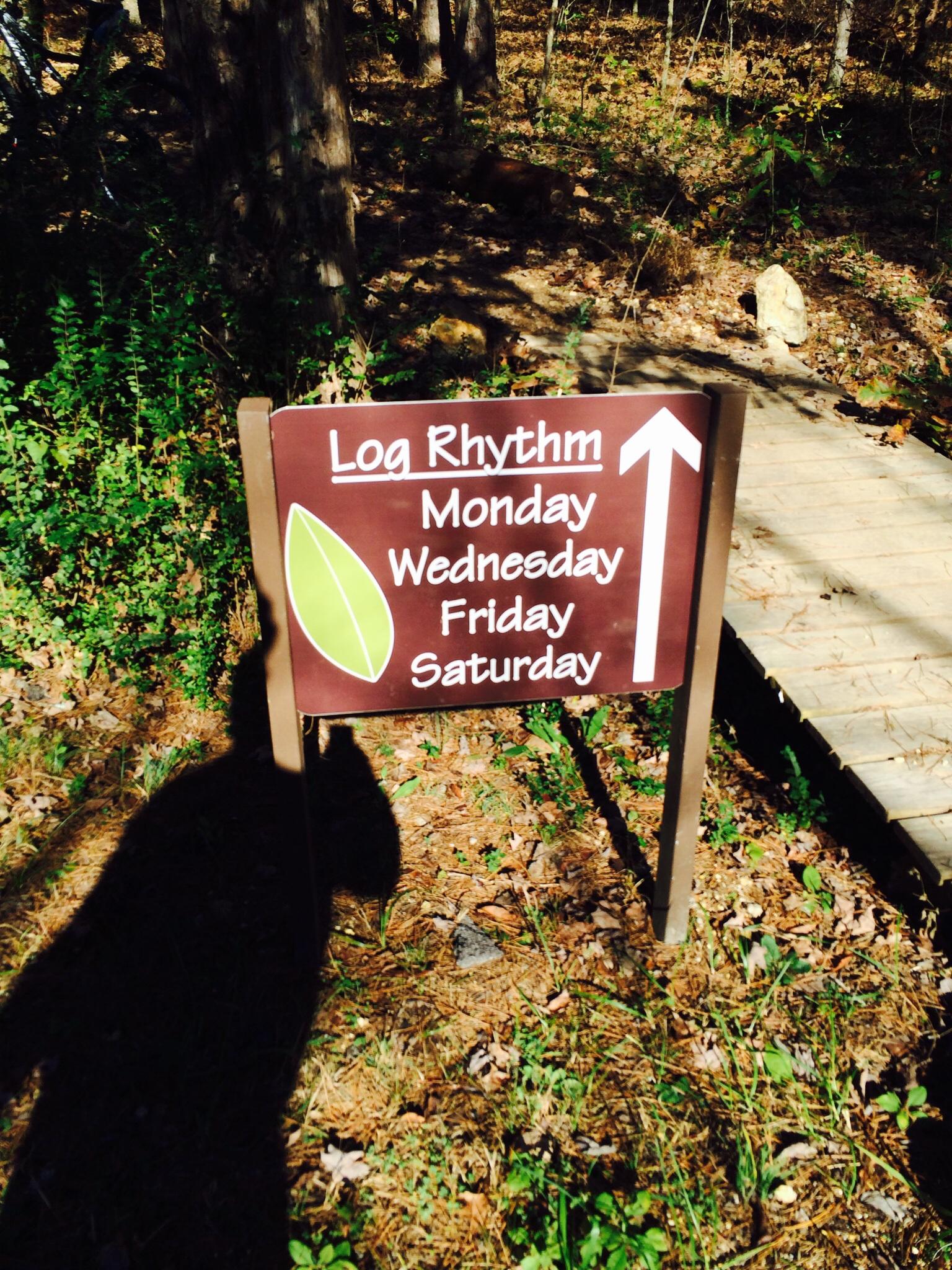 A brown wooden sign in a forested area displaying "Log Rhythm" along with the days of the week: Monday, Wednesday, Friday, and Saturday. An arrow pointing upward indicates the direction to follow. The background is filled with trees and greenery. Enterprise South mountain bike trail.