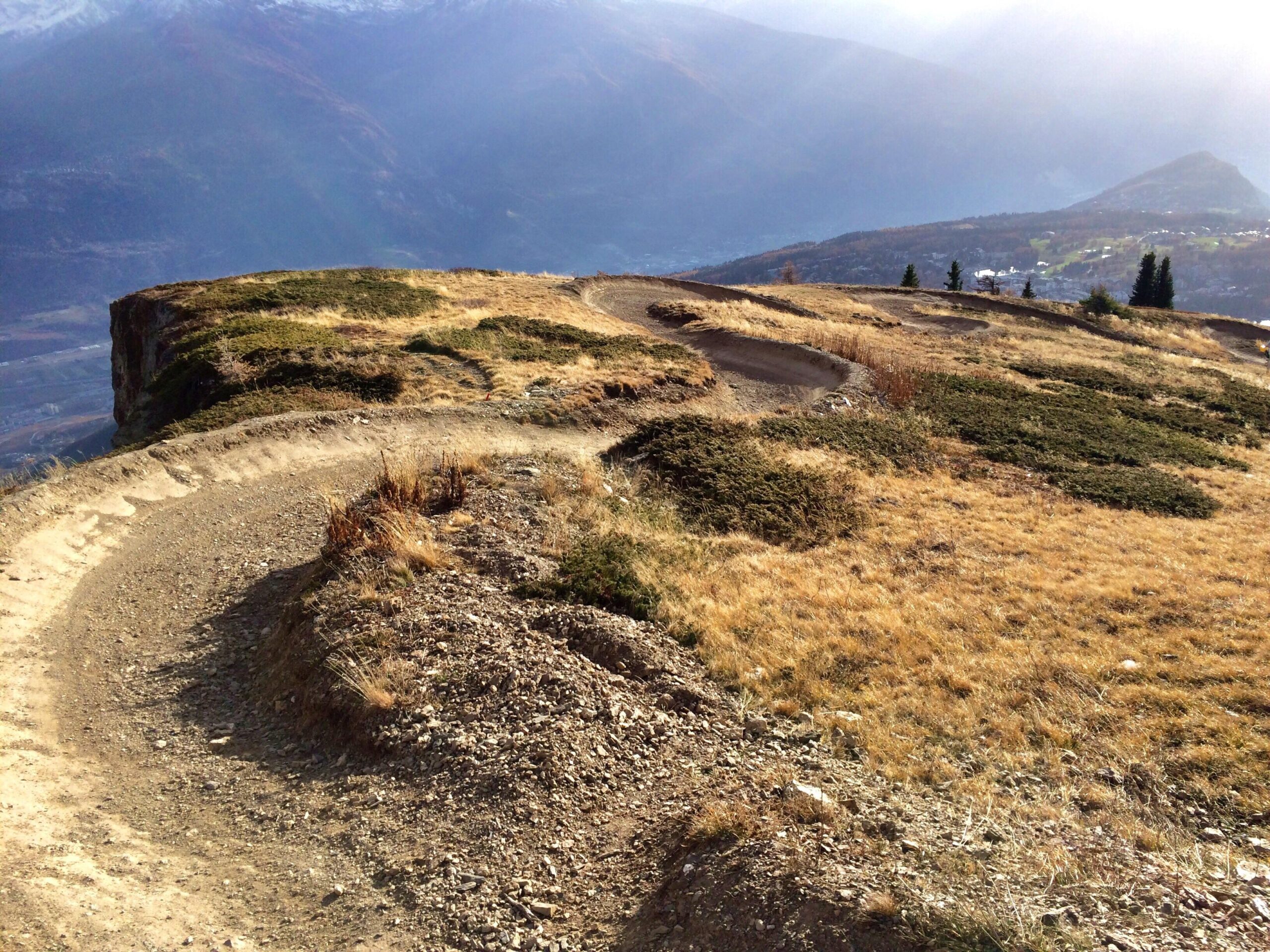 A winding dirt path cuts through a landscape of dry grass and rocky terrain, leading to a steep cliff edge with distant mountains visible in the background. The scene is bathed in soft sunlight, highlighting the natural beauty of the area. Cran-montana mountain bike trail.