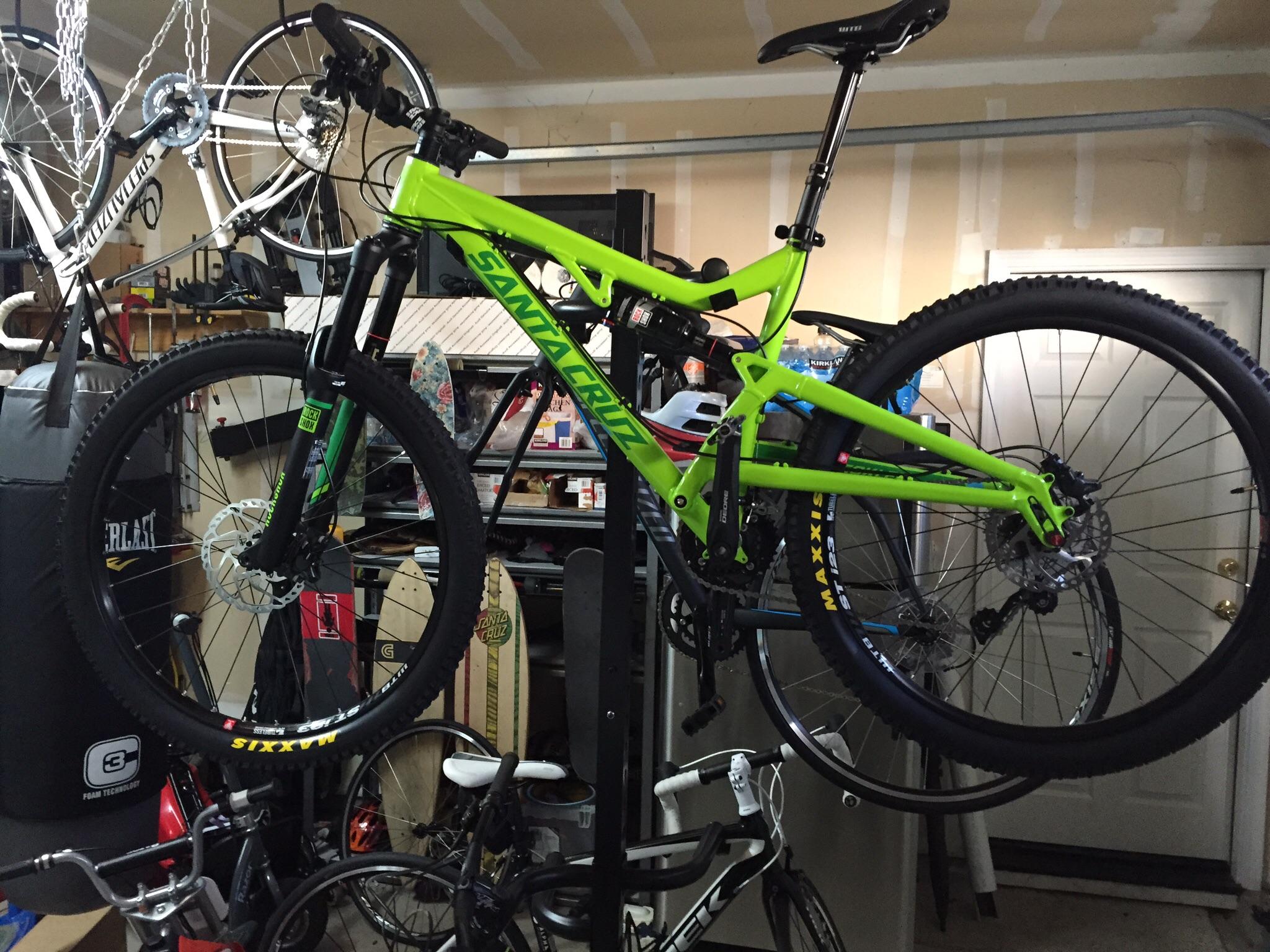 Santa Cruz Heckler: A bright green mountain bike hanging from a ceiling rack in a garage, with additional bicycles and a punching bag visible in the background. The garage has unfinished drywall and various sporting equipment arranged around the space.