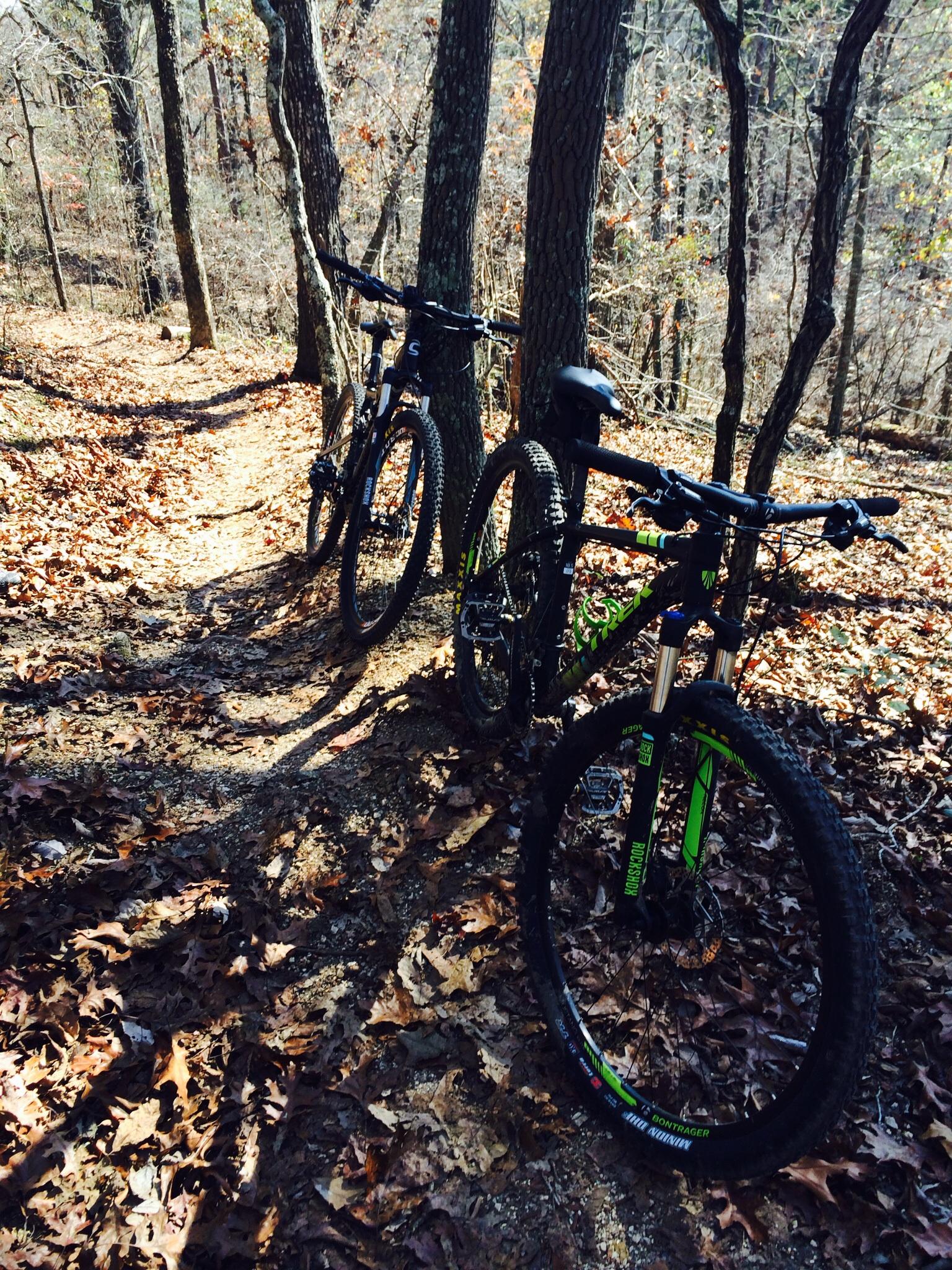 Two mountain bikes parked beside a winding dirt trail surrounded by trees in a wooded area, with fallen leaves covering the ground. Enterprise South mountain bike trail.