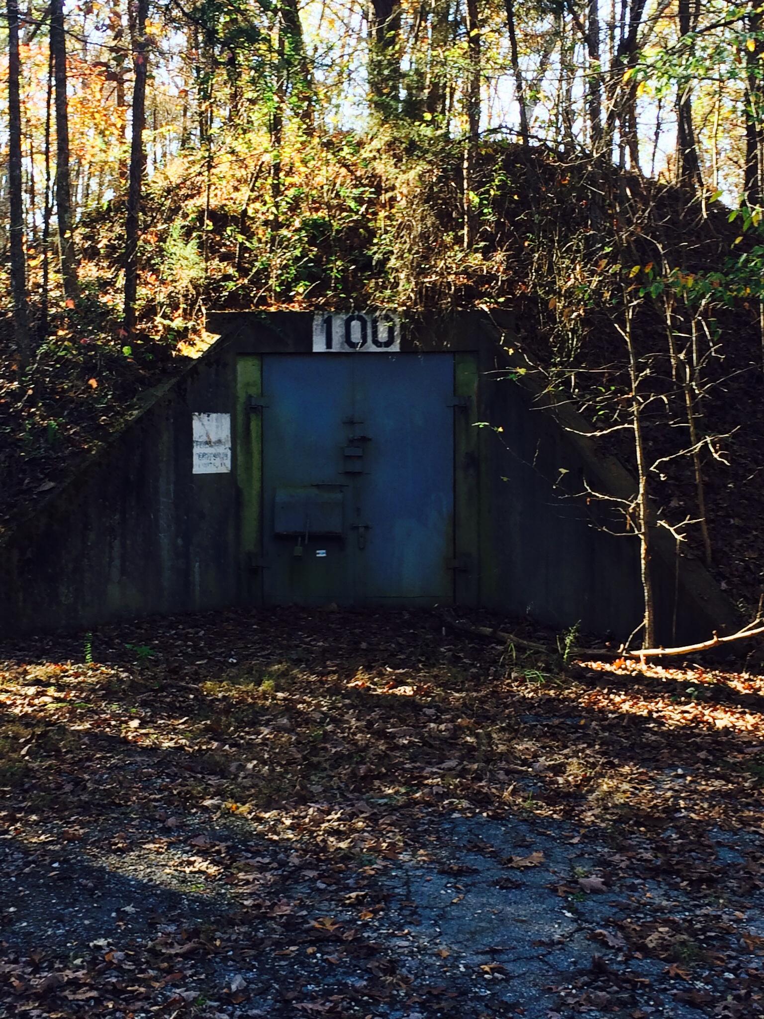 A weathered green door marked with the number 100, set into a hillside surrounded by trees displaying autumn foliage. The area is covered in fallen leaves, with hints of sunlight filtering through the trees. Enterprise South mountain bike trail.